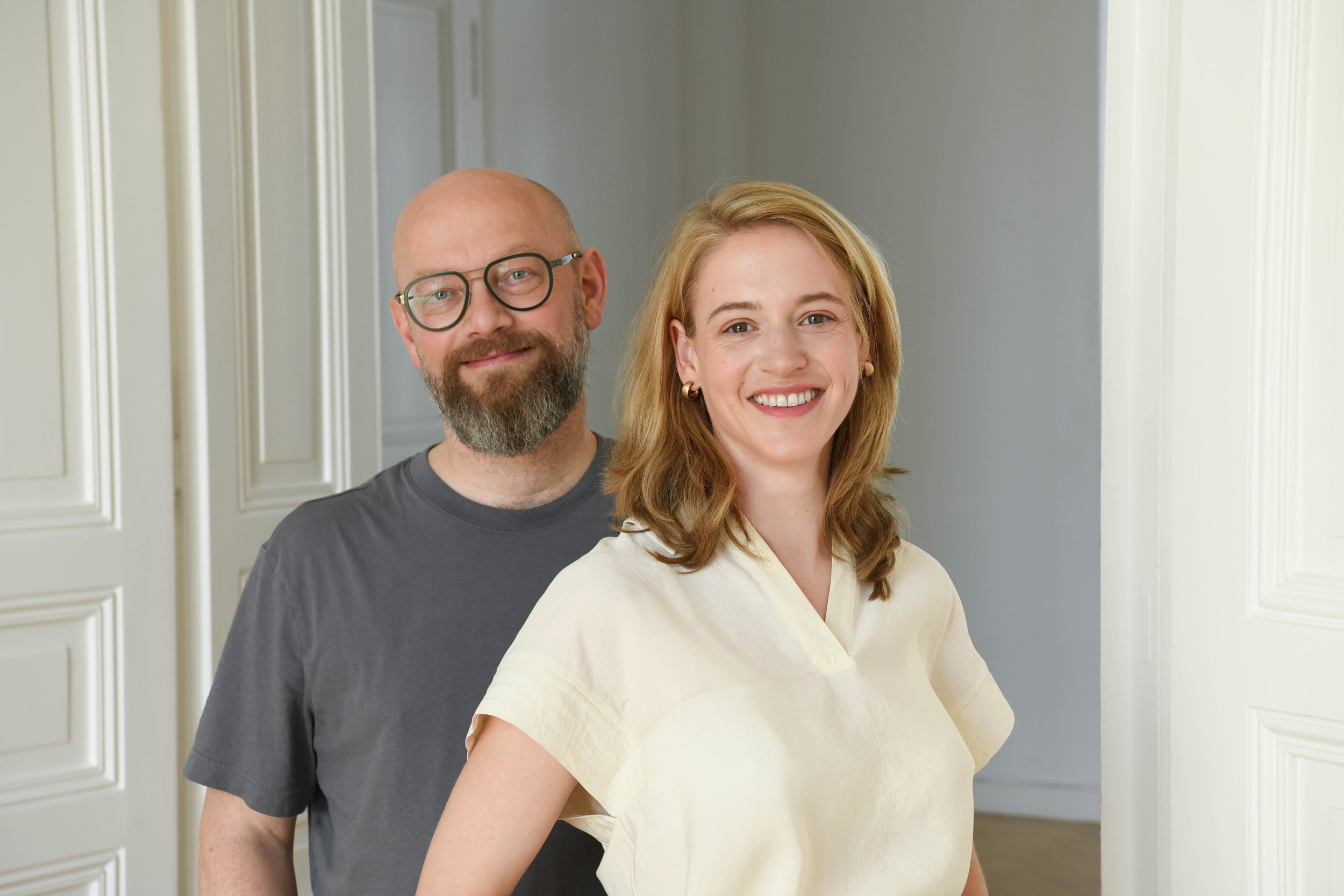 Ein lächelndes Mann und eine Frau stehen vor einer weißen Wand mit Türrahmen im Hintergrund.