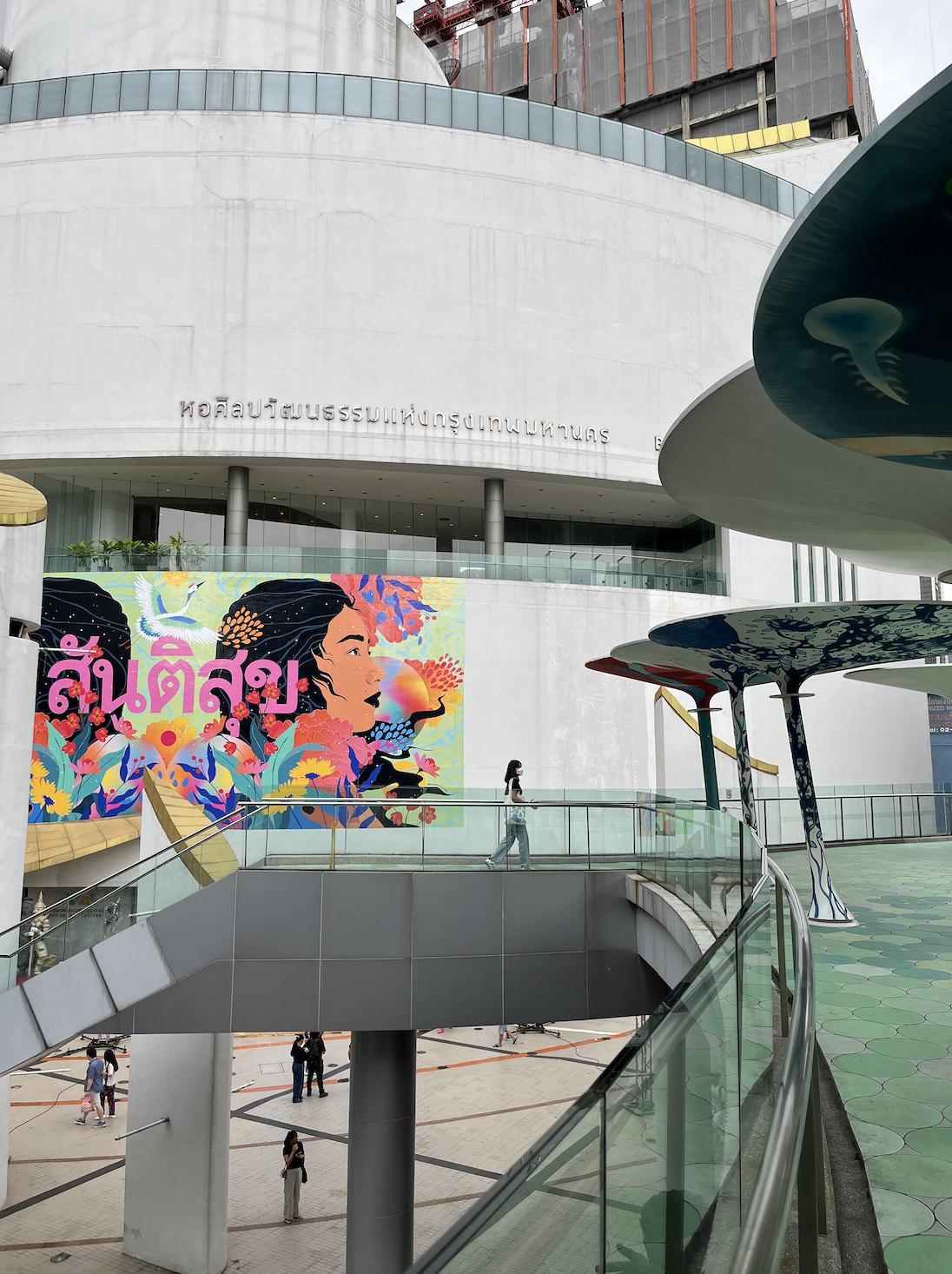 Modern building with a colorful mural of a woman's profile, tropical birds, and flowers on the wall, with a person walking on a glass walkway and large mushroom-shaped structures in the foreground.