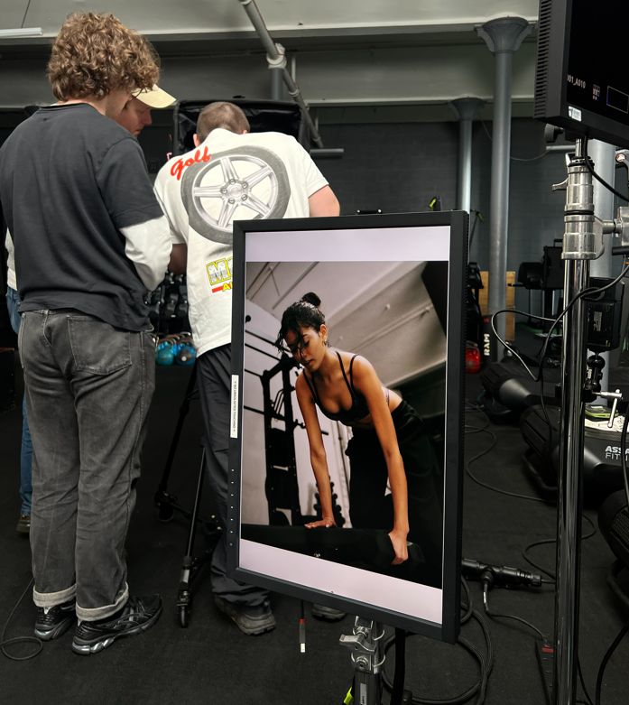 A photoshoot scene with a model performing an exercise, viewed on a screen in front of the camera crew.