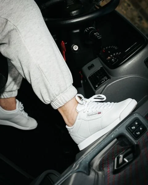 Person wearing white Reebok sneakers with their foot on the car's dashboard while sitting in the driver's seat.