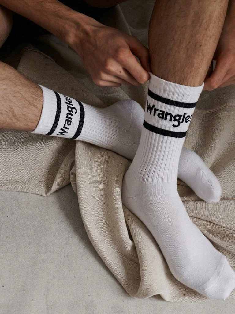 A person wearing white socks with black stripes and the brand name 'Wrangler,' adjusting the sock of another person's foot. The setting appears to be indoors on a beige fabric surface.