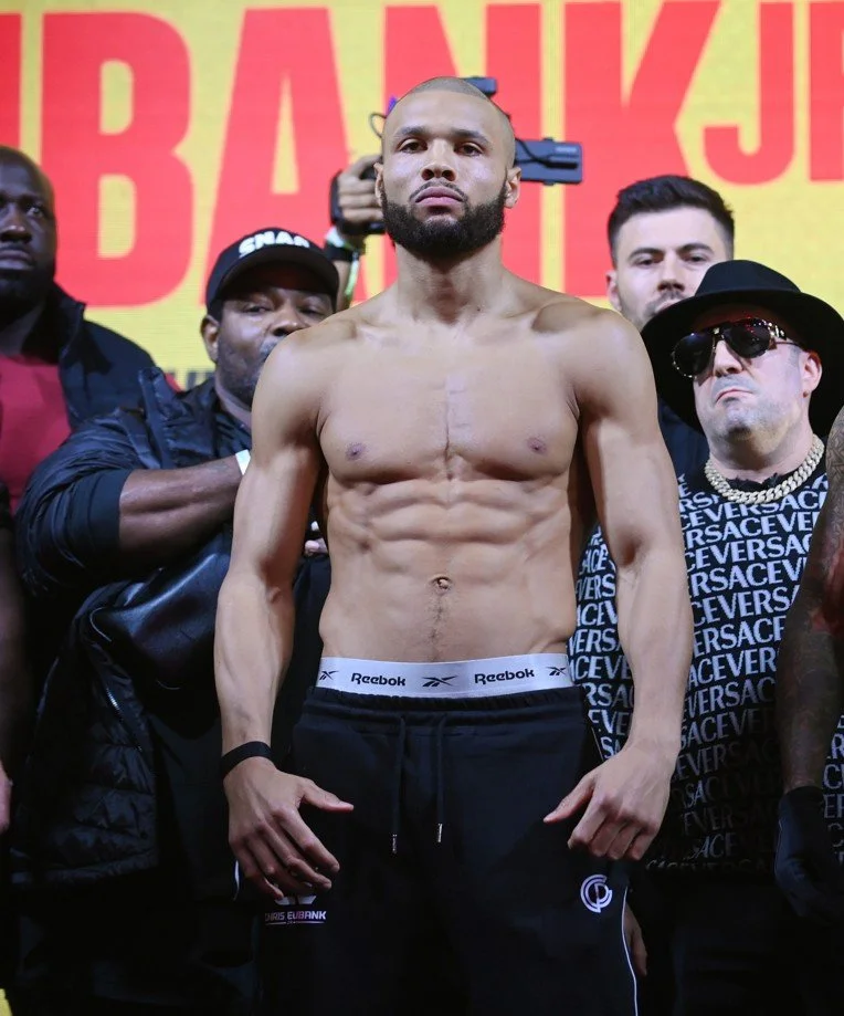 A shirtless male boxer with a beard and muscular build standing on a weigh-in stage, surrounded by several other men. The background has large yellow and red text.