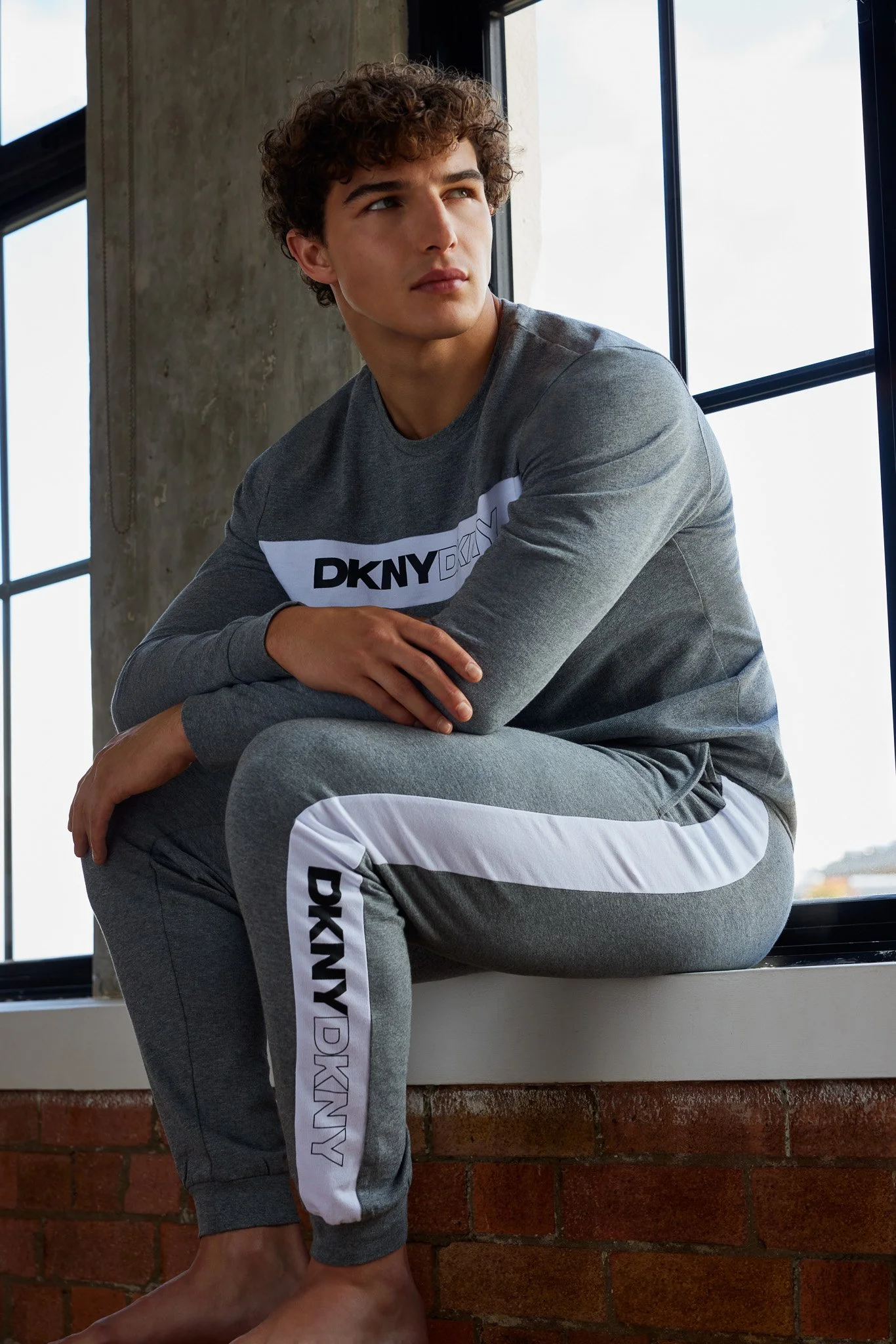 A young man with curly brown hair sitting on a windowsill, looking thoughtfully to the side, wearing a gray DKNY sweatshirt and matching sweatpants with white Nike side stripes, near a brick wall.