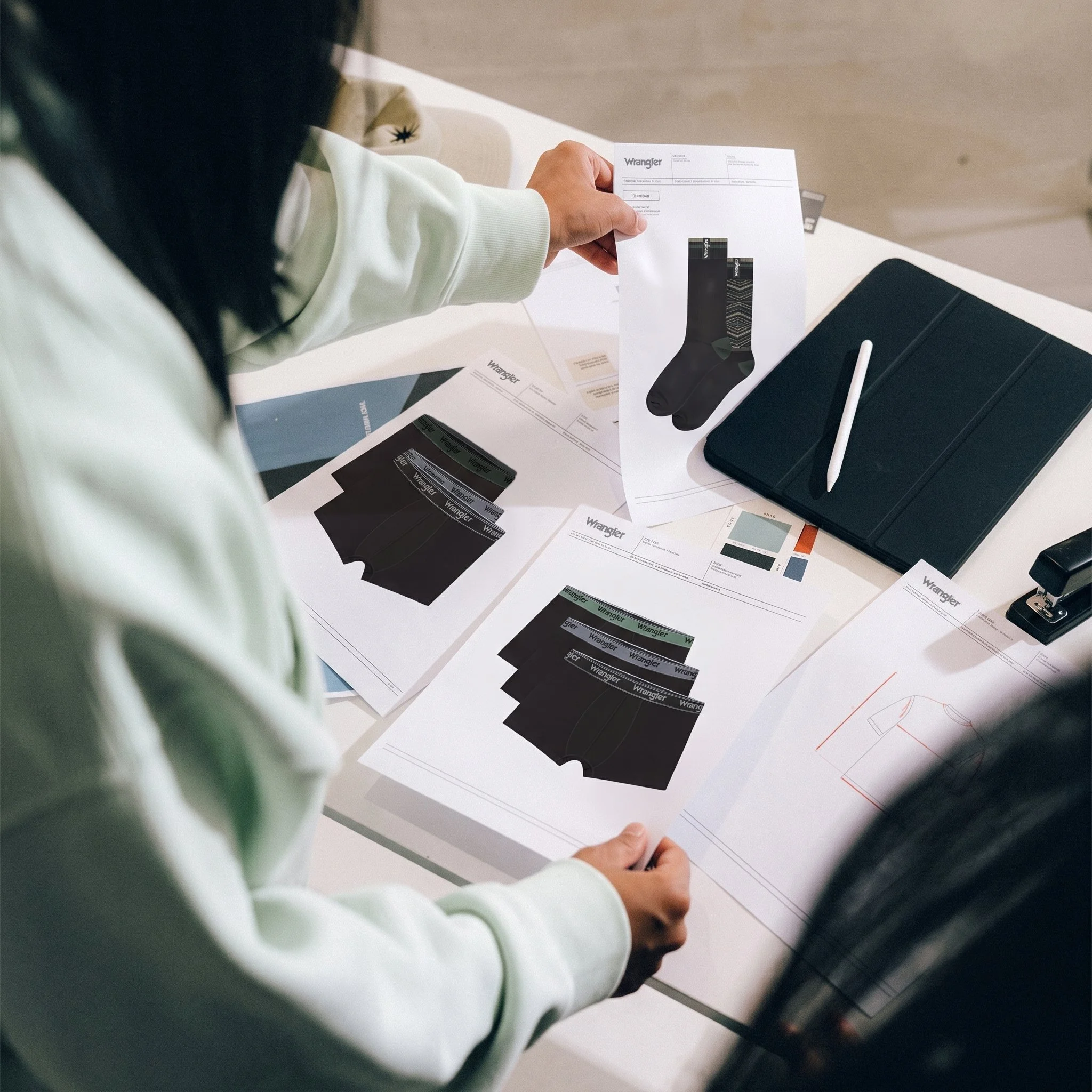 Person holding a white paper with a black sock design printed on it, surrounded by clothing and office supplies, on a white desk.