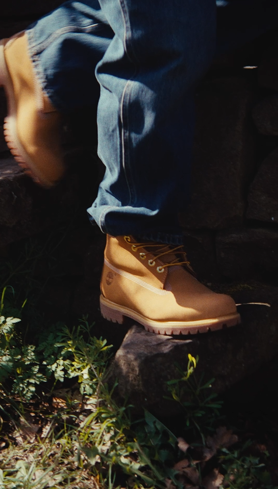 Close-up of a person wearing tan work boots and blue jeans, stepping on a stone in a natural outdoor setting with greenery.