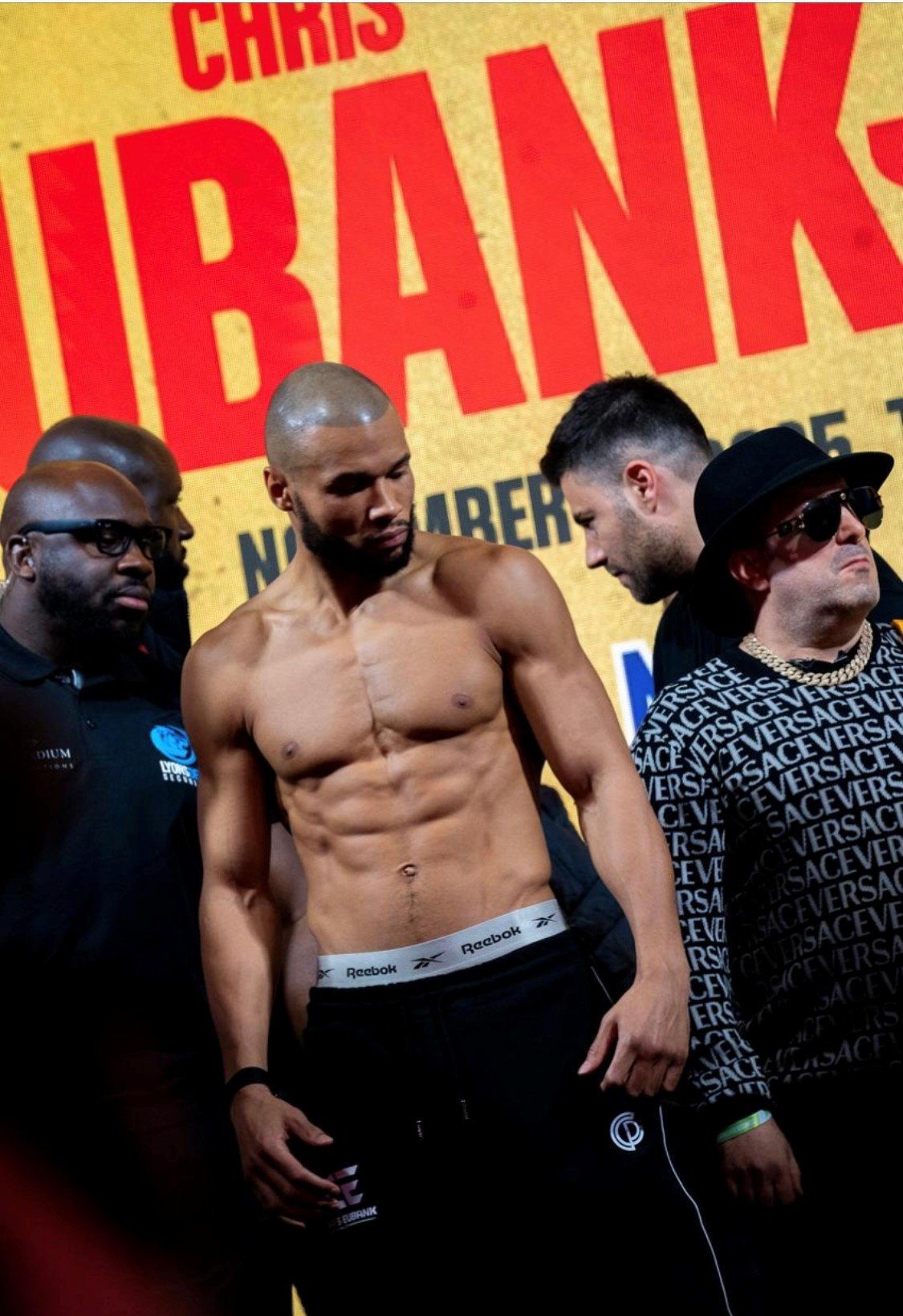 A shirtless male boxer on a weighing scale during a weigh-in event, with several men around him, one wearing sunglasses, a black hat, and a Versace-patterned shirt, against a yellow background with red text that reads 'CHRIS IBRAHIM'.