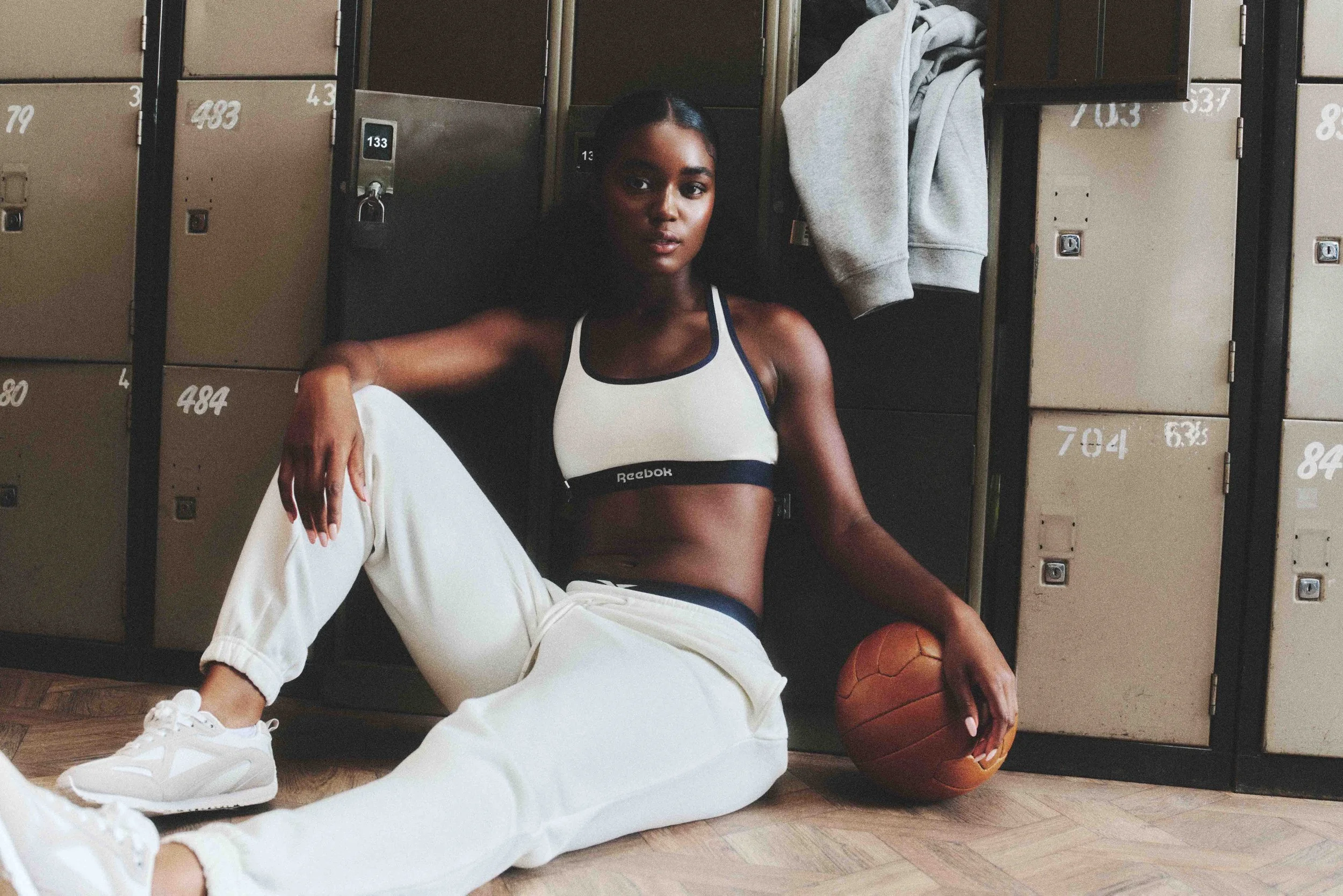 A fit woman in athletic wear sitting on the floor in a locker room, holding a basketball, with wooden lockers in the background.