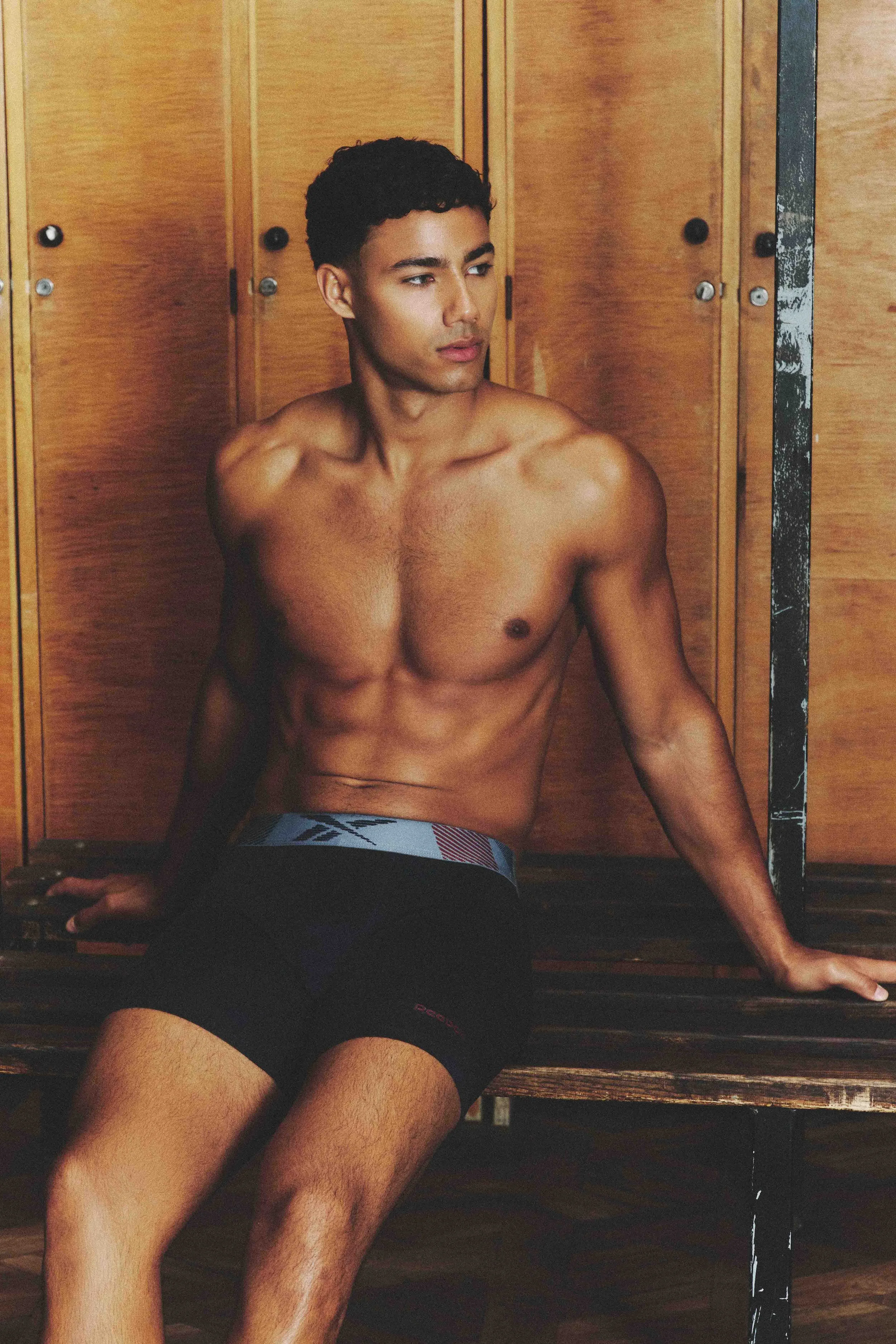 A shirtless young man with dark curly hair and toned physique sitting on a wooden bench in front of wooden lockers.