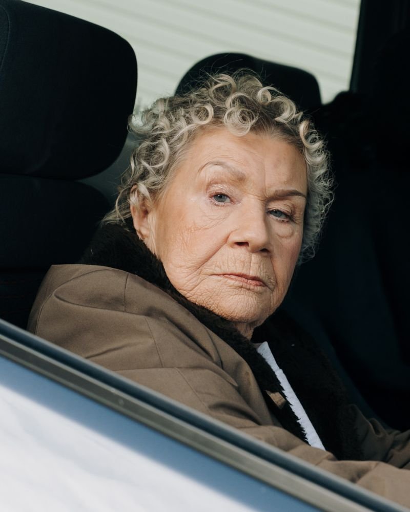An elderly woman with curly gray hair looking out of a car window with a serious expression.