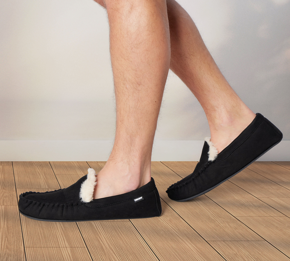 A person wearing black slippers with white fur lining, standing on a wooden floor.