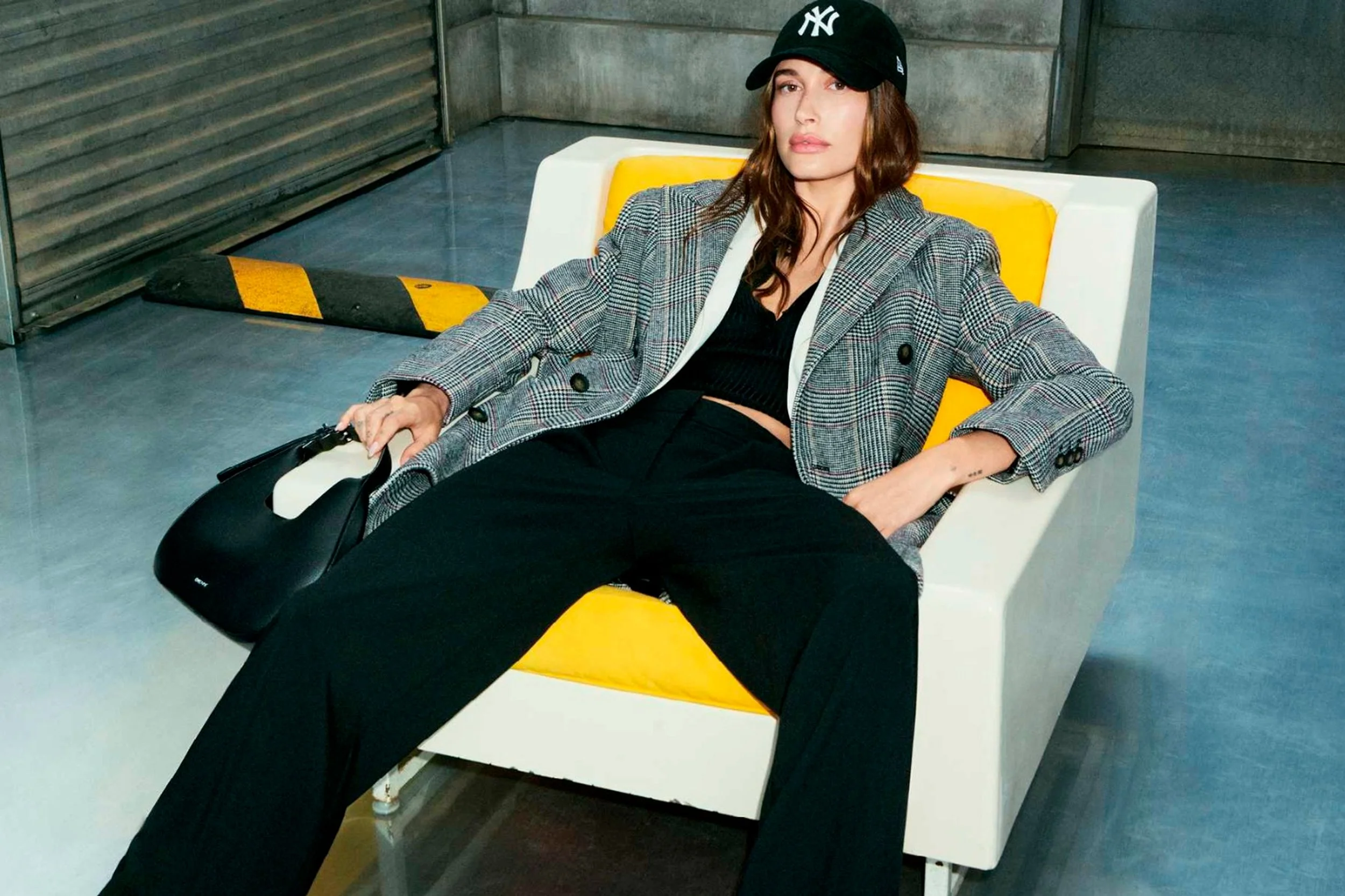 A woman sitting behind a yellow and white barrier, wearing a black NY cap, plaid blazer, black top, and black pants, with one arm resting on her leg and holding a black bag.