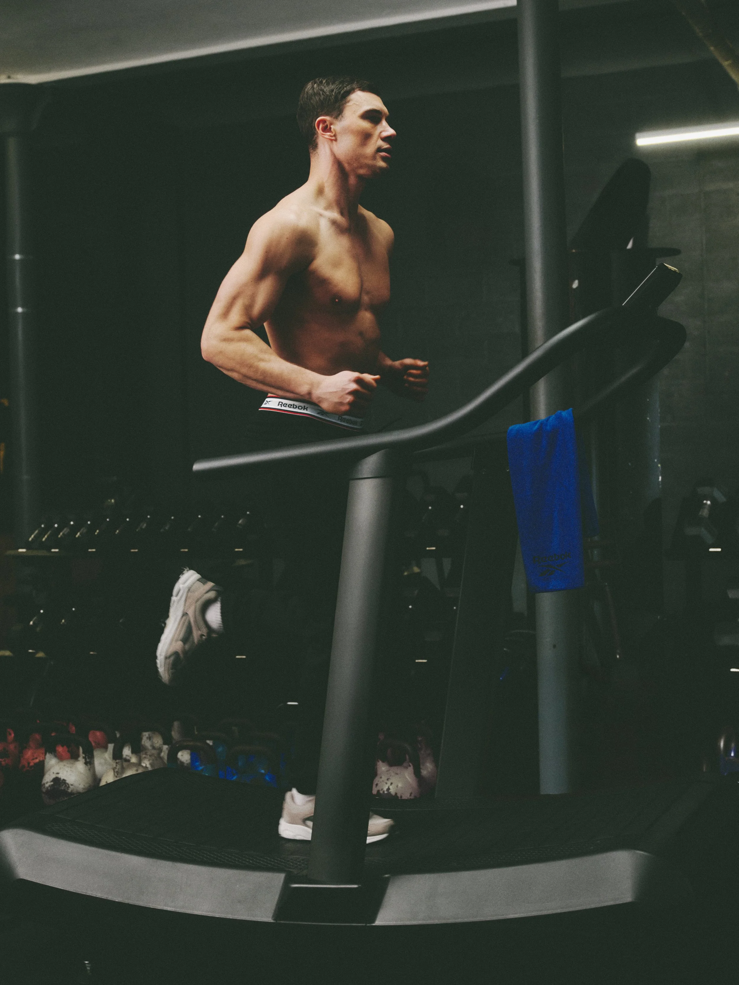 A shirtless man running on a treadmill in a gym, with gym equipment and weights in the background.