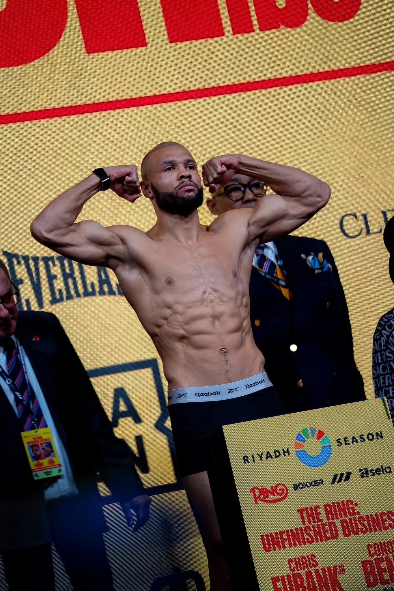 A shirtless man with a muscular build, beard, and shaved head, holding his arms up to show his biceps, standing in front of a yellow background with red text. Two men in suits are standing behind him, one wearing glasses and a tie, the other with a lanyard and badge, on an event stage.