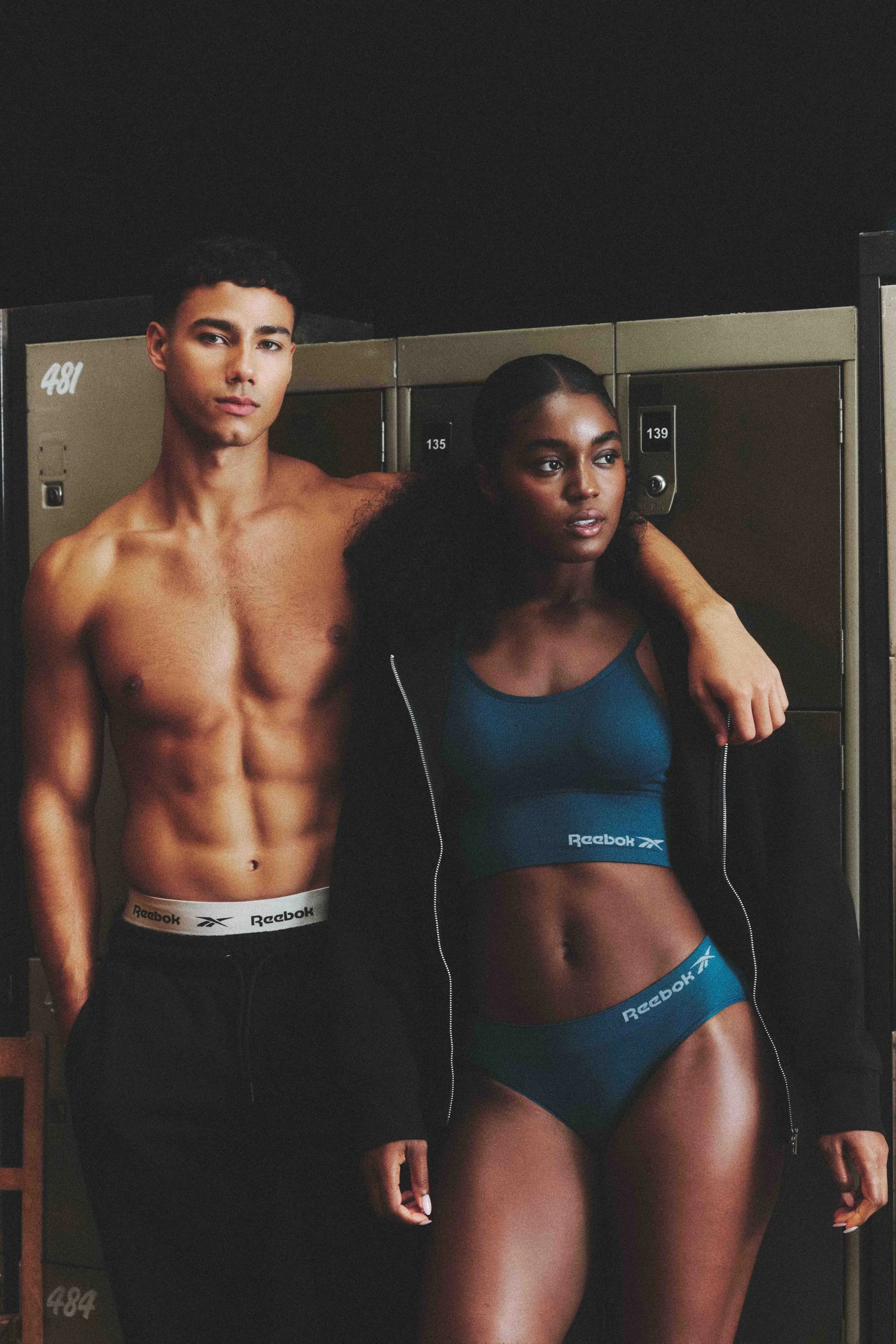 A shirtless man and a woman in athletic wear are standing together in front of lockers in a gym, looking at the camera.