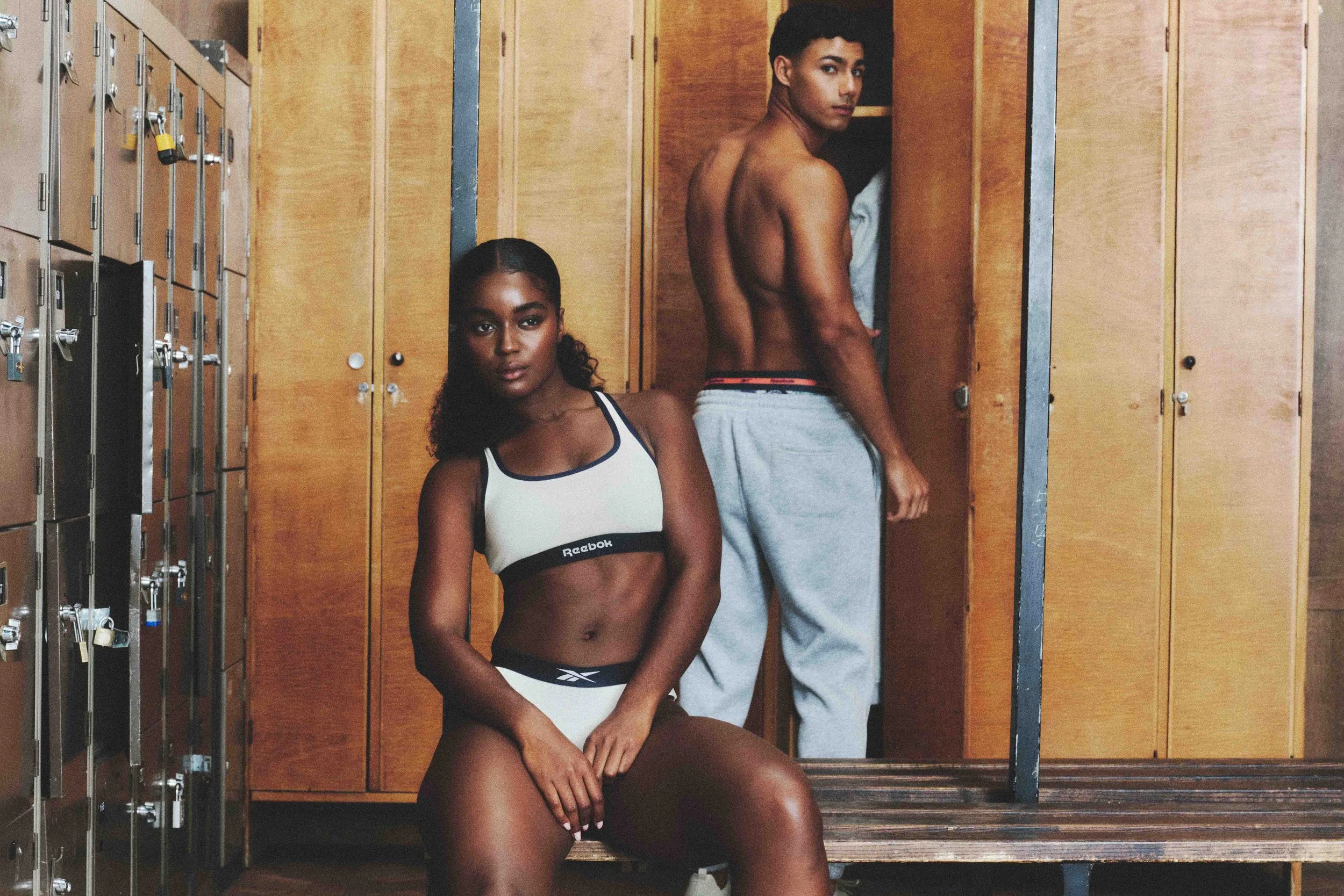 A woman in sports bra and shorts sitting on a bench in a locker room, with a shirtless man standing behind her, looking over his shoulder.