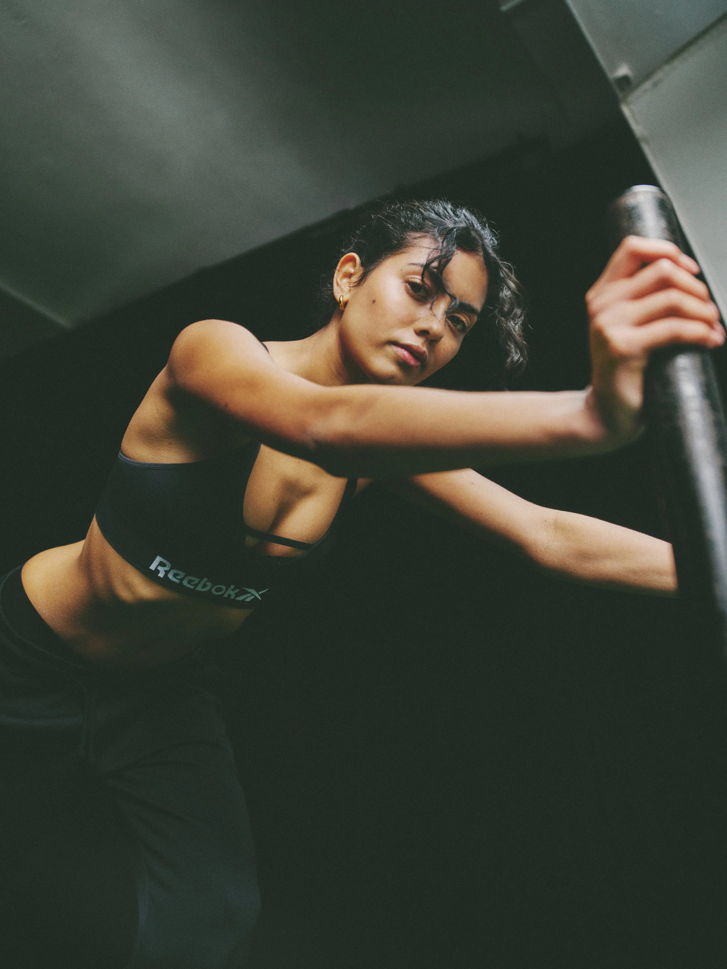A woman with dark curly hair in workout attire stretching in a gym setting.