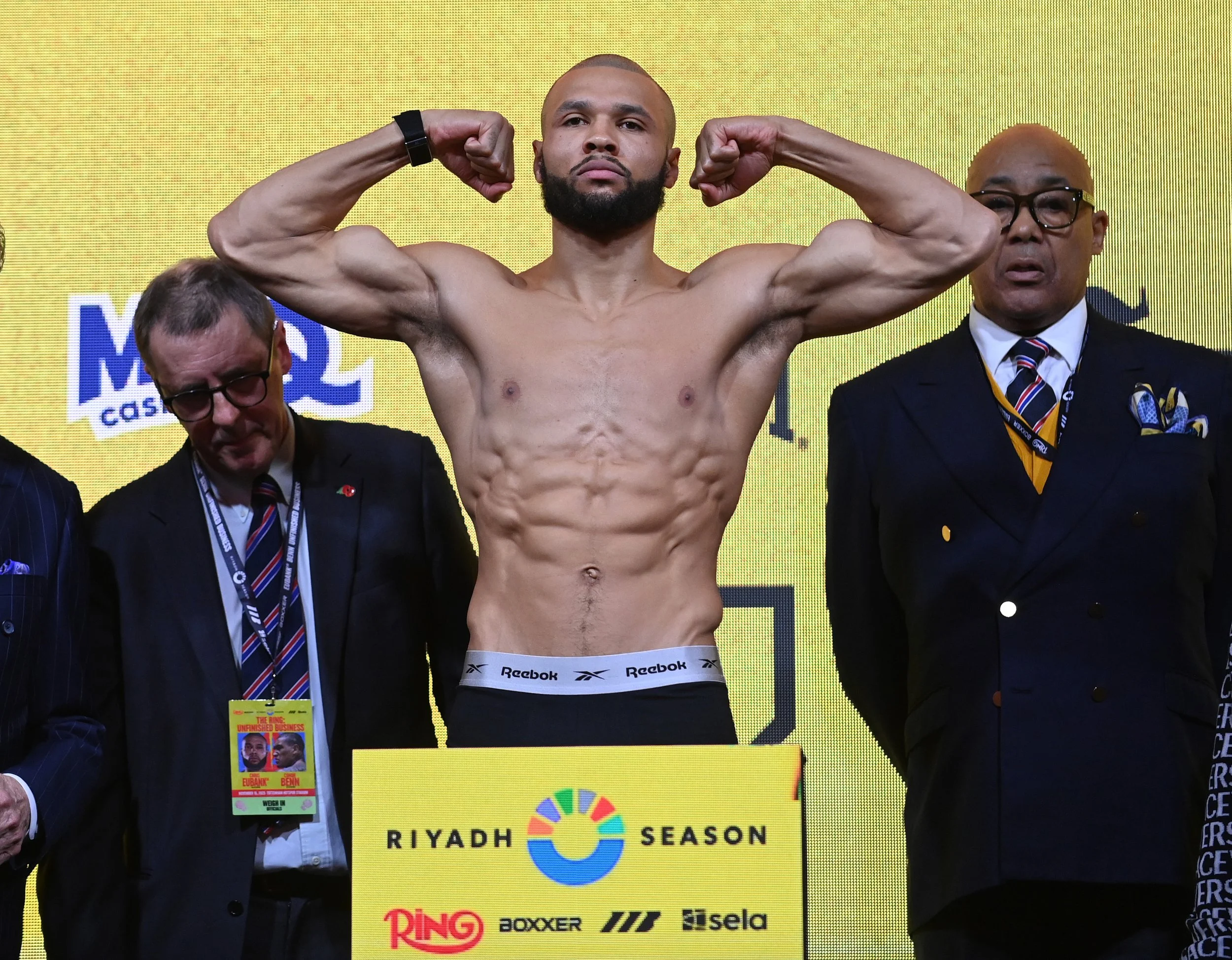 A male boxer with a muscular build and a beard, standing on a stage with his arms raised in a victory pose. He is shirtless, wearing black shorts with the Reebok logo. Two men in suits are standing beside him, one on each side. The background is yellow with text and logos, including