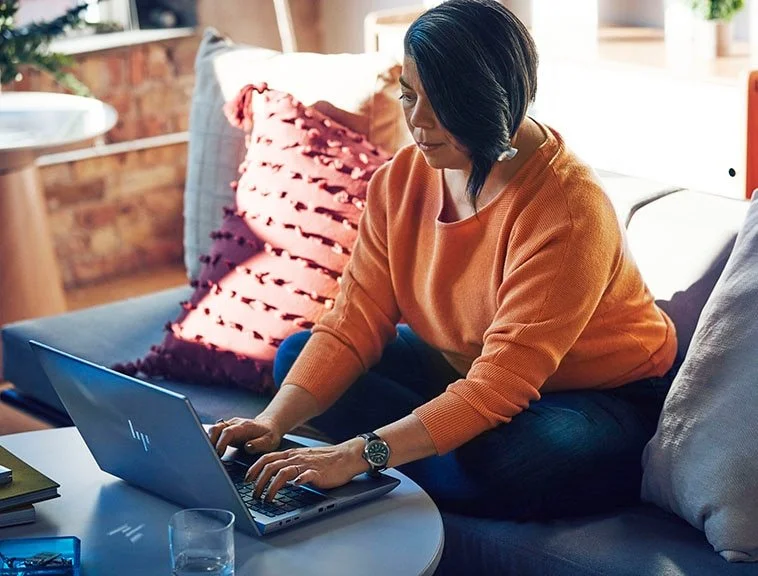 A woman in an orange sweater working on a silver HP laptop in a cozy living room with cushions and natural light.