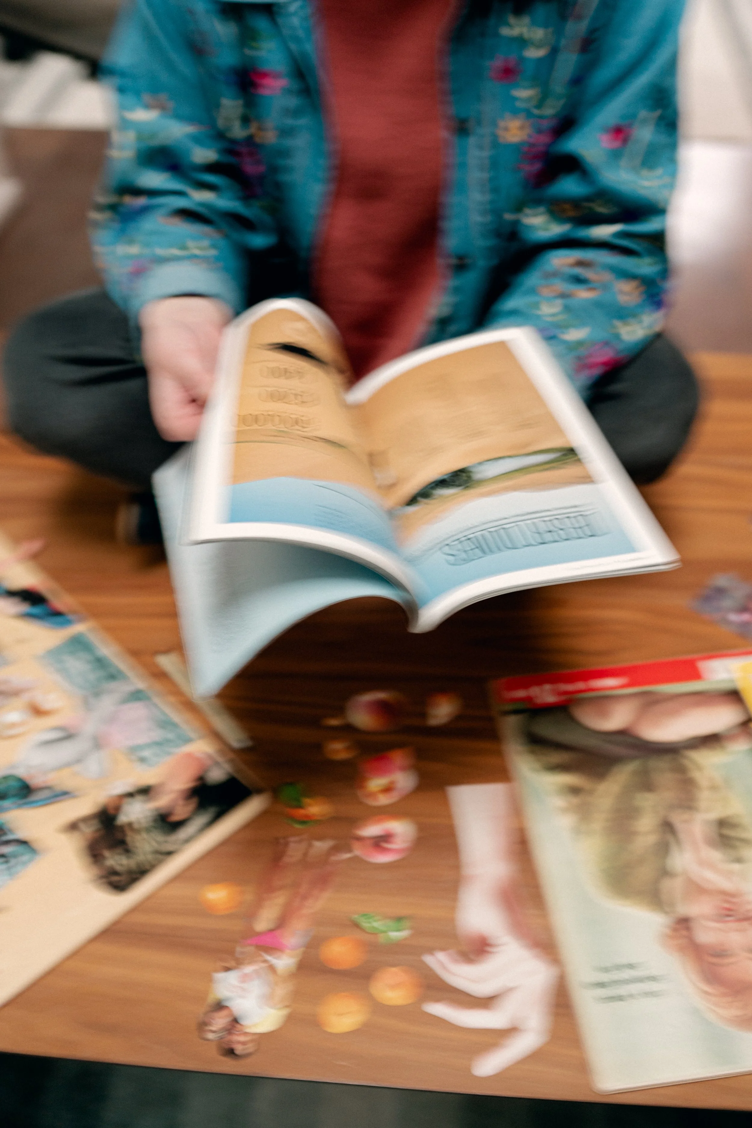 Blurred image of a person sitting at a table reading a booklet or magazine, surrounded by other magazines and scattered small colorful objects.