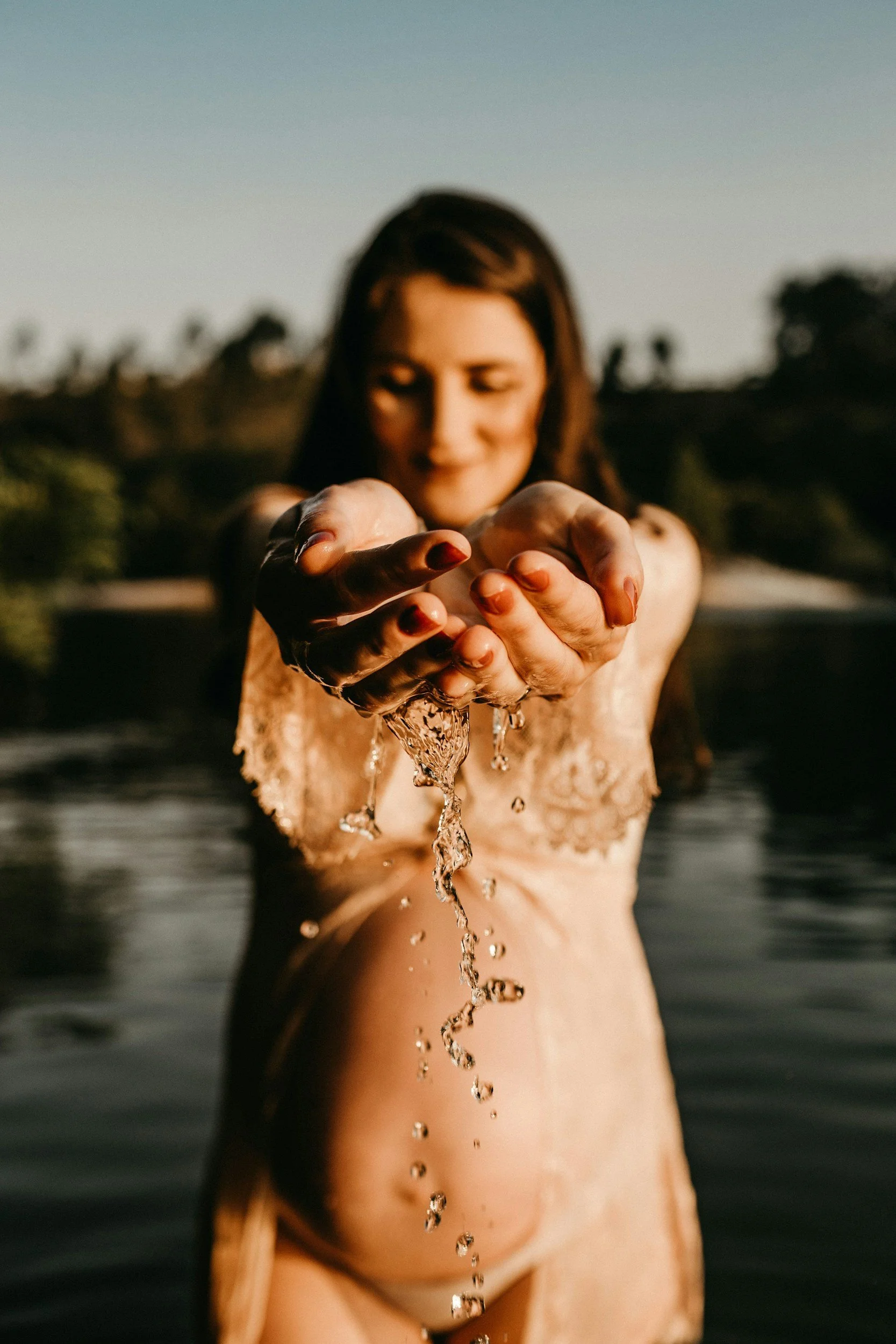 Eine Frau steht im Wasser und hält ihre Hände nach vorne, wobei Wasser über ihre Finger läuft. Der Hintergrund zeigt einen klaren Himmel und eine ländliche Landschaft.