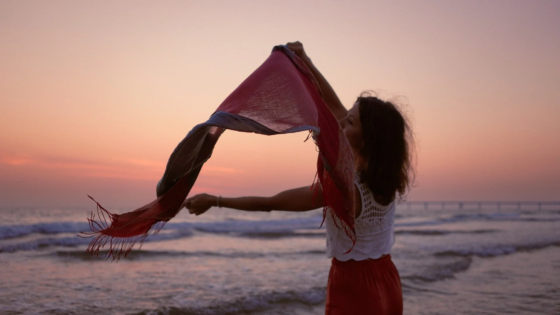 Éloïse tenant une écharpe rouge face à la mer au coucher du soleil