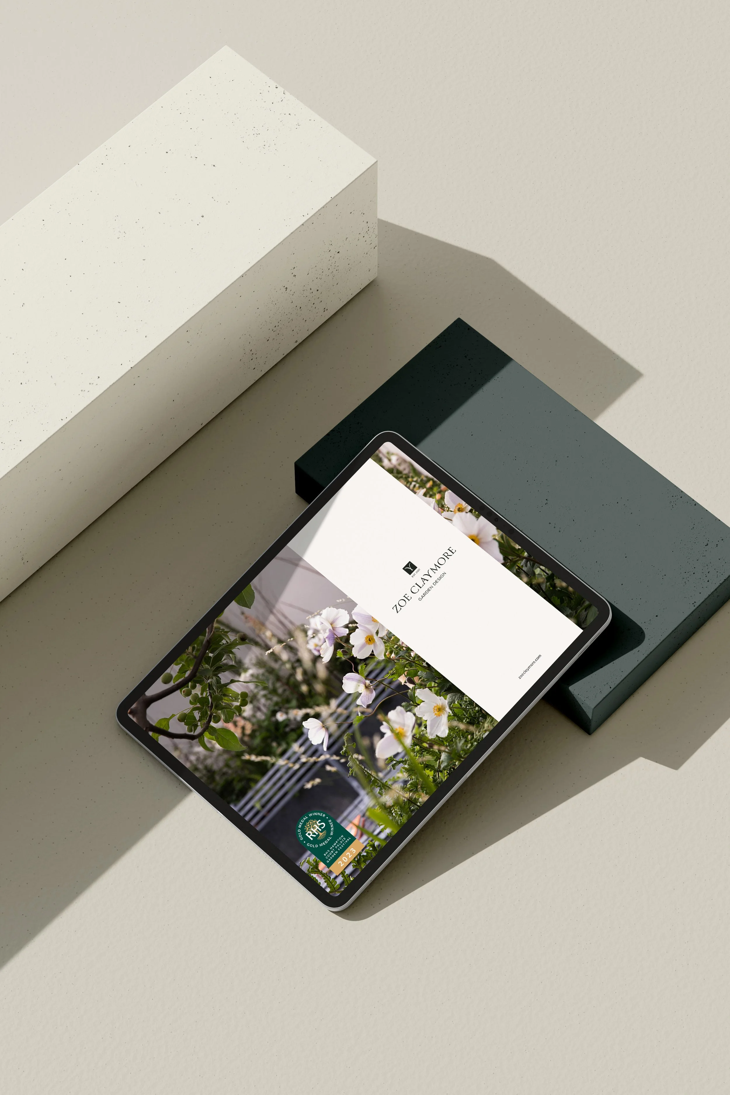 A smartphone displaying a gardening website with an image of white flowers and a gold medal badge for 2023. The phone rests on a light-colored surface beside geometric blocks.