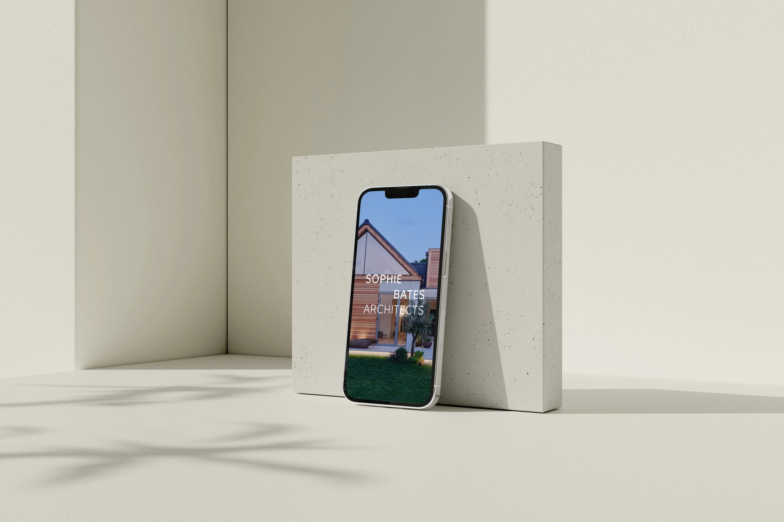 A smartphone leaning against a gray, textured block displaying a house and the text "Sophie Bates Architects" on the screen.
