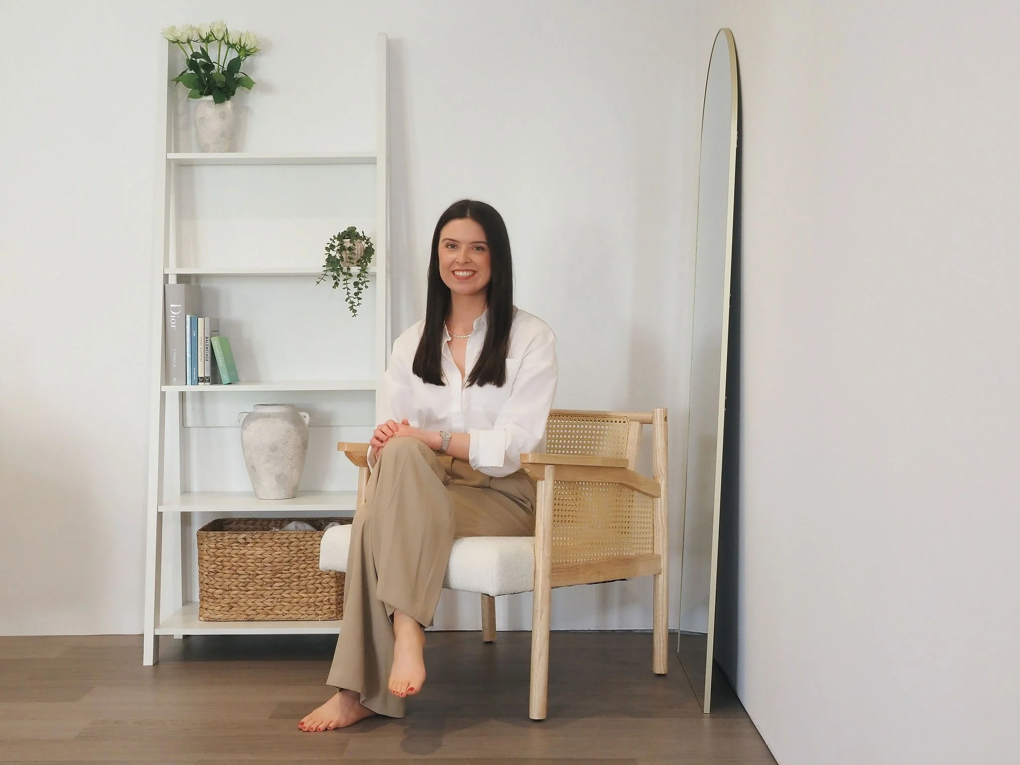 A woman with long dark hair smiling, seated on a wooden chair in a minimalist room with white walls, next to a white shelf with books and plants, and a large mirror.