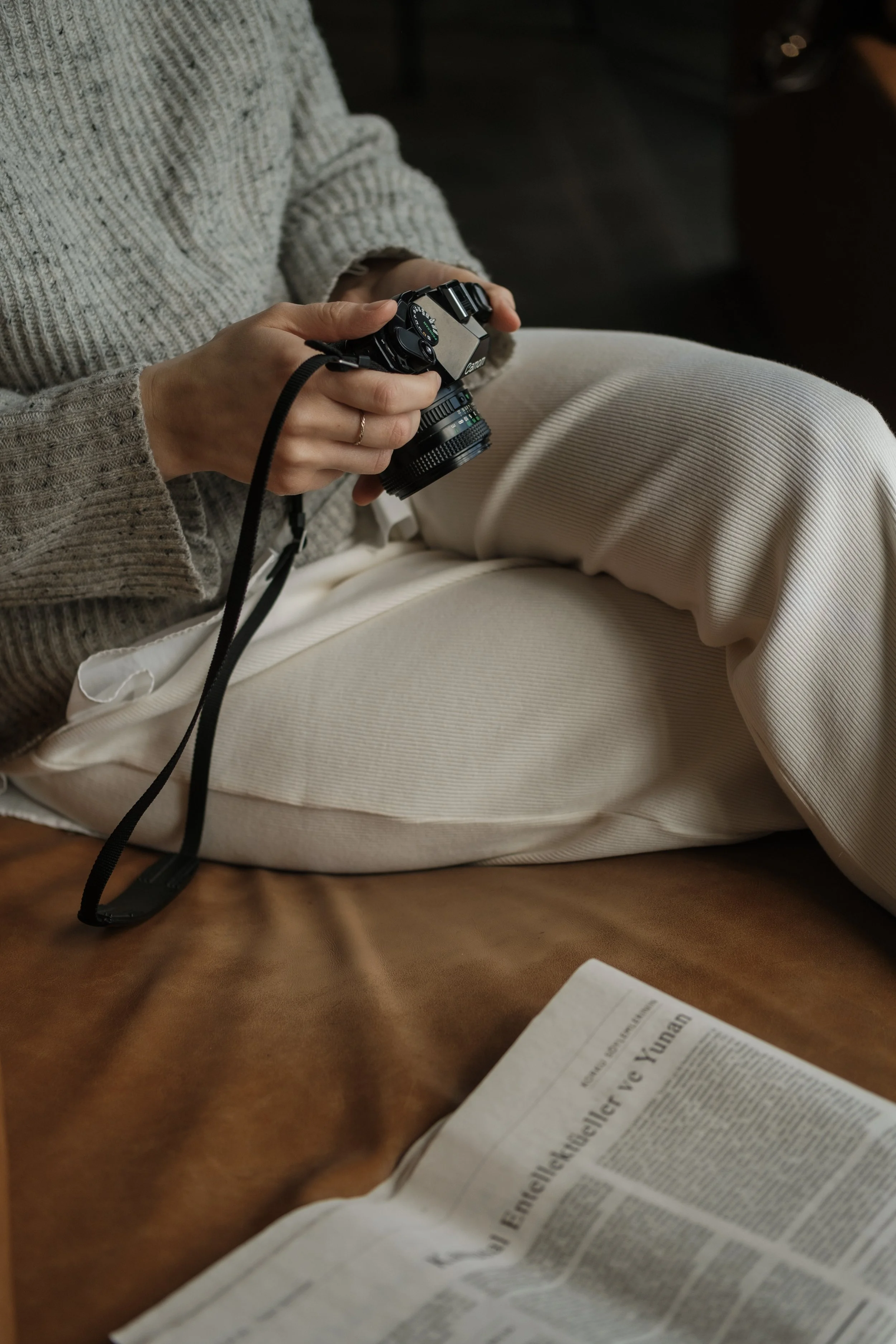A person sitting on a brown leather surface, holding a Canon camera with both hands, wearing a beige sweater and light-colored pants, with a newspaper on their lap.