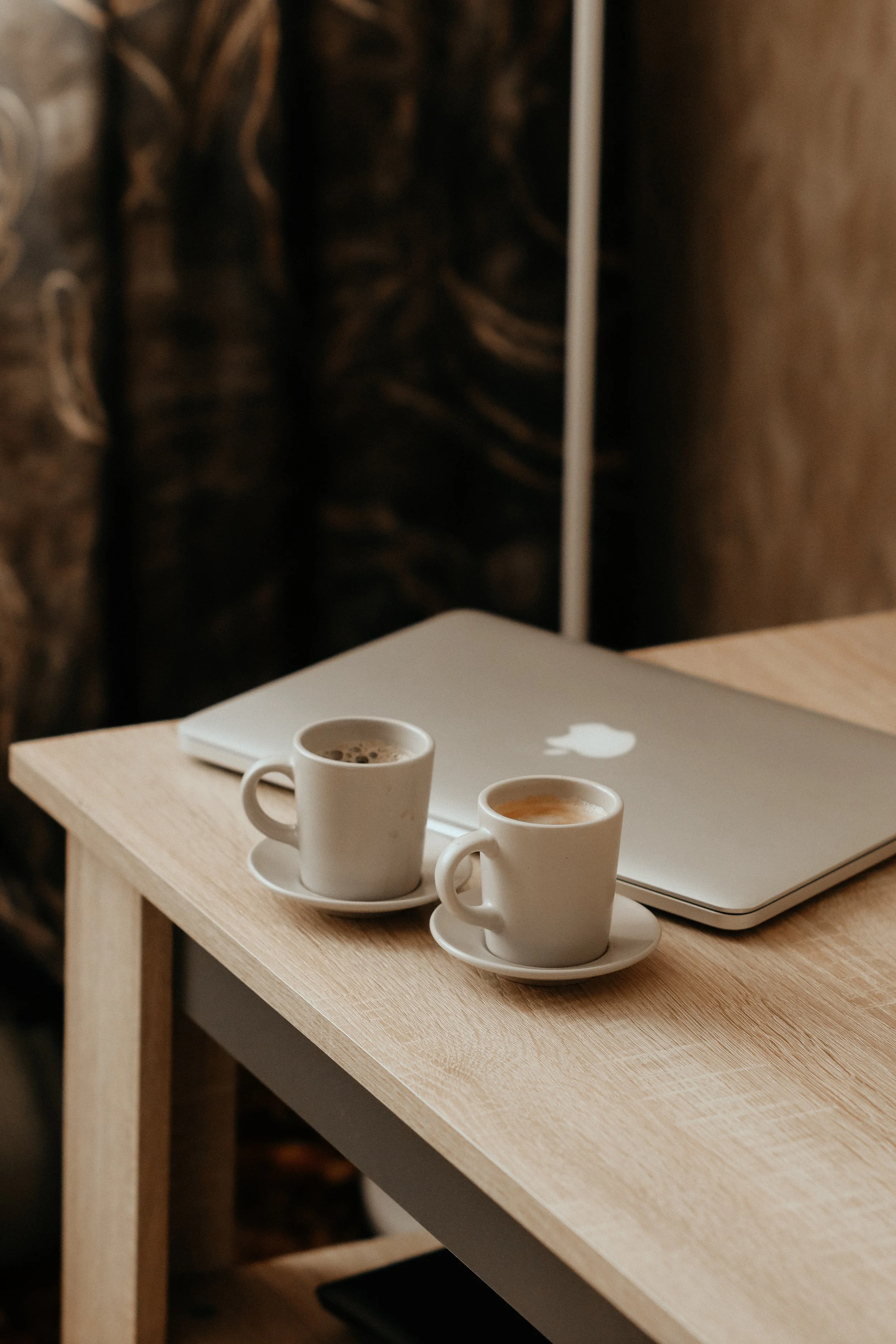 Two white coffee cups with saucers, a silver MacBook laptop, and an open coffee bag are on a light wooden desk.