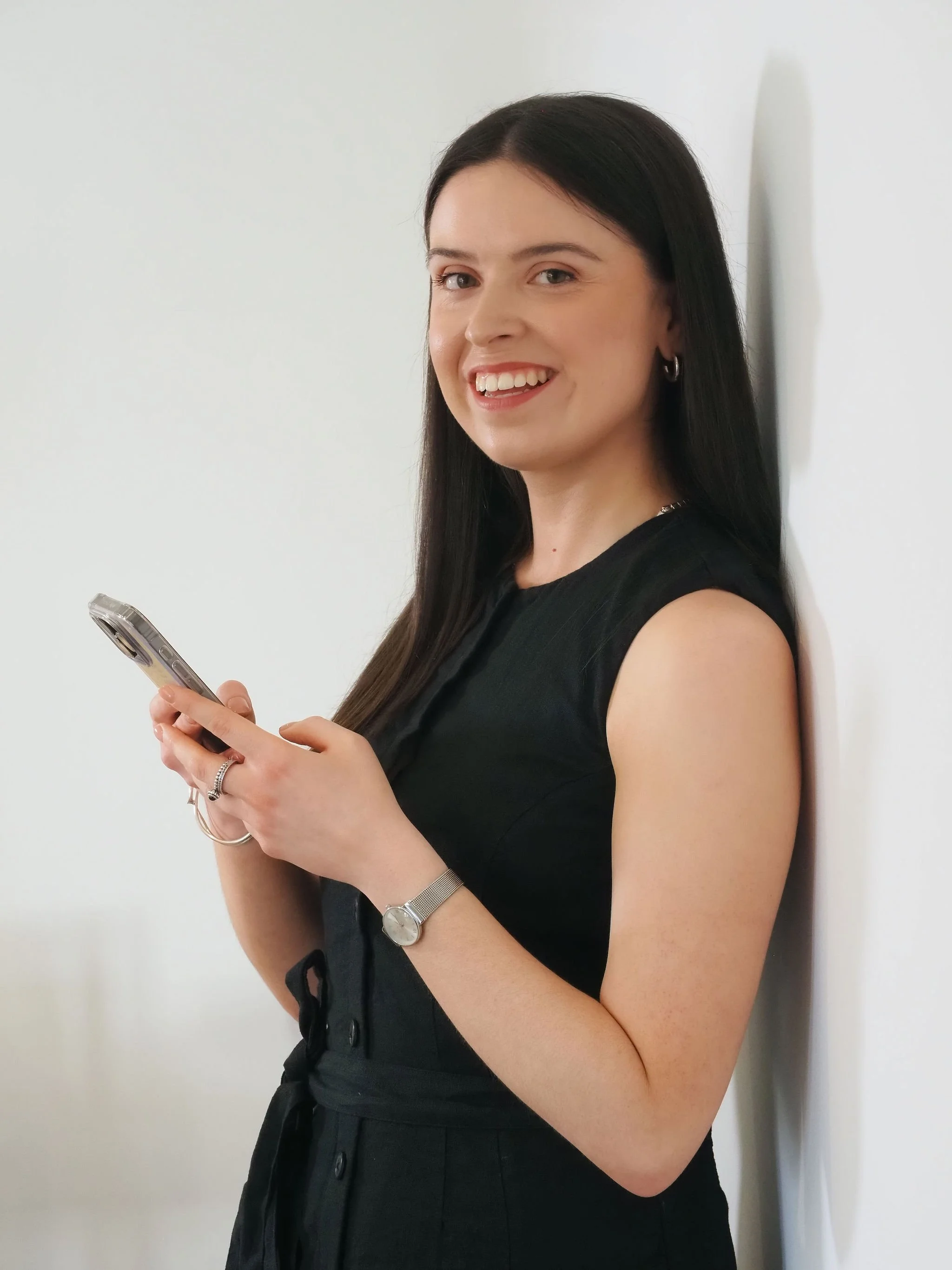 A woman with long dark hair and hoop earrings, wearing a black sleeveless dress, is smiling while looking at her smartphone. She is standing against a white wall.