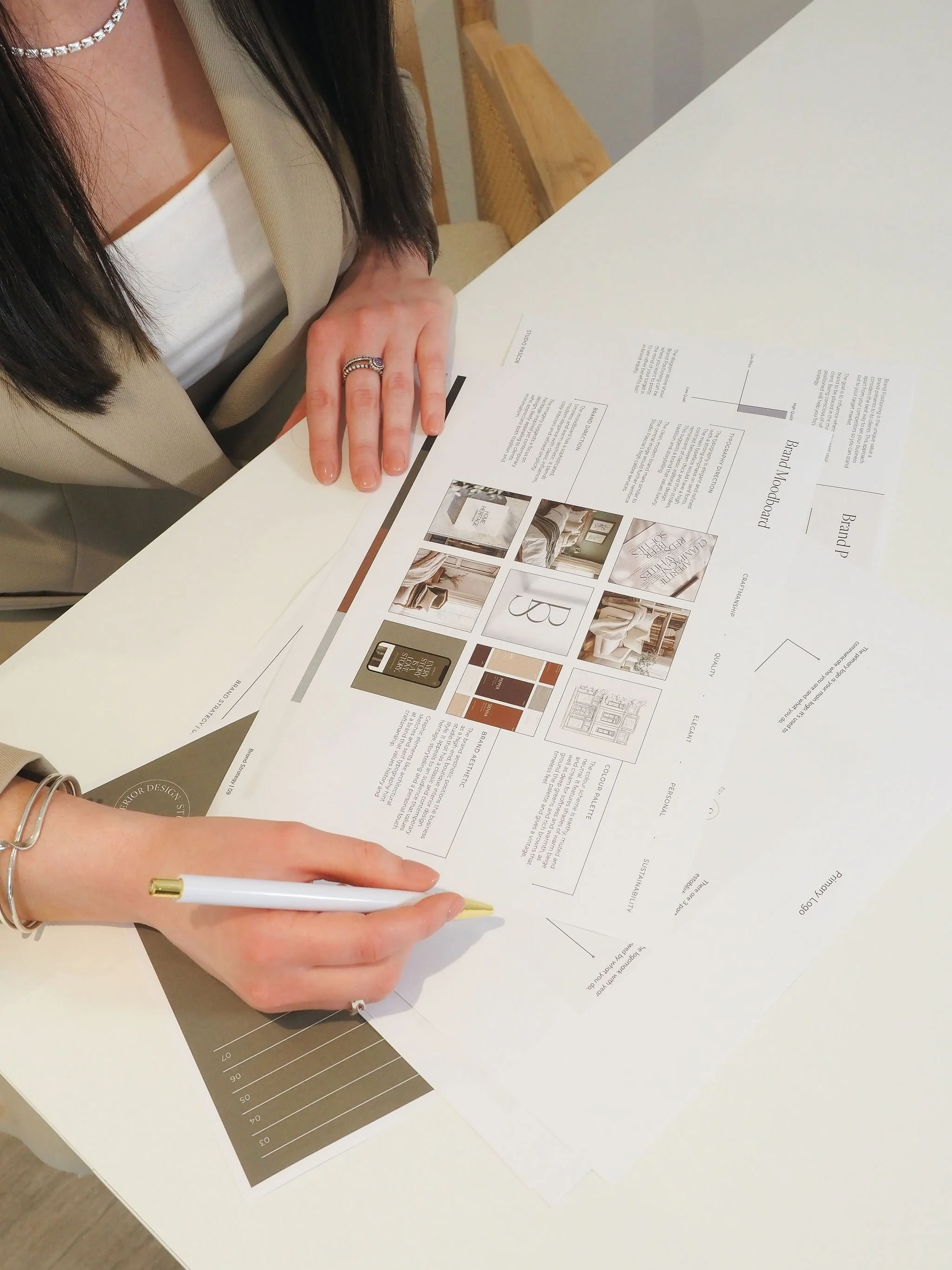 A woman wearing a beige blazer and white top, with a pearl necklace and rings, is pointing with a white pen at a paper on a white table. The paper contains images and text about interior design and branding.