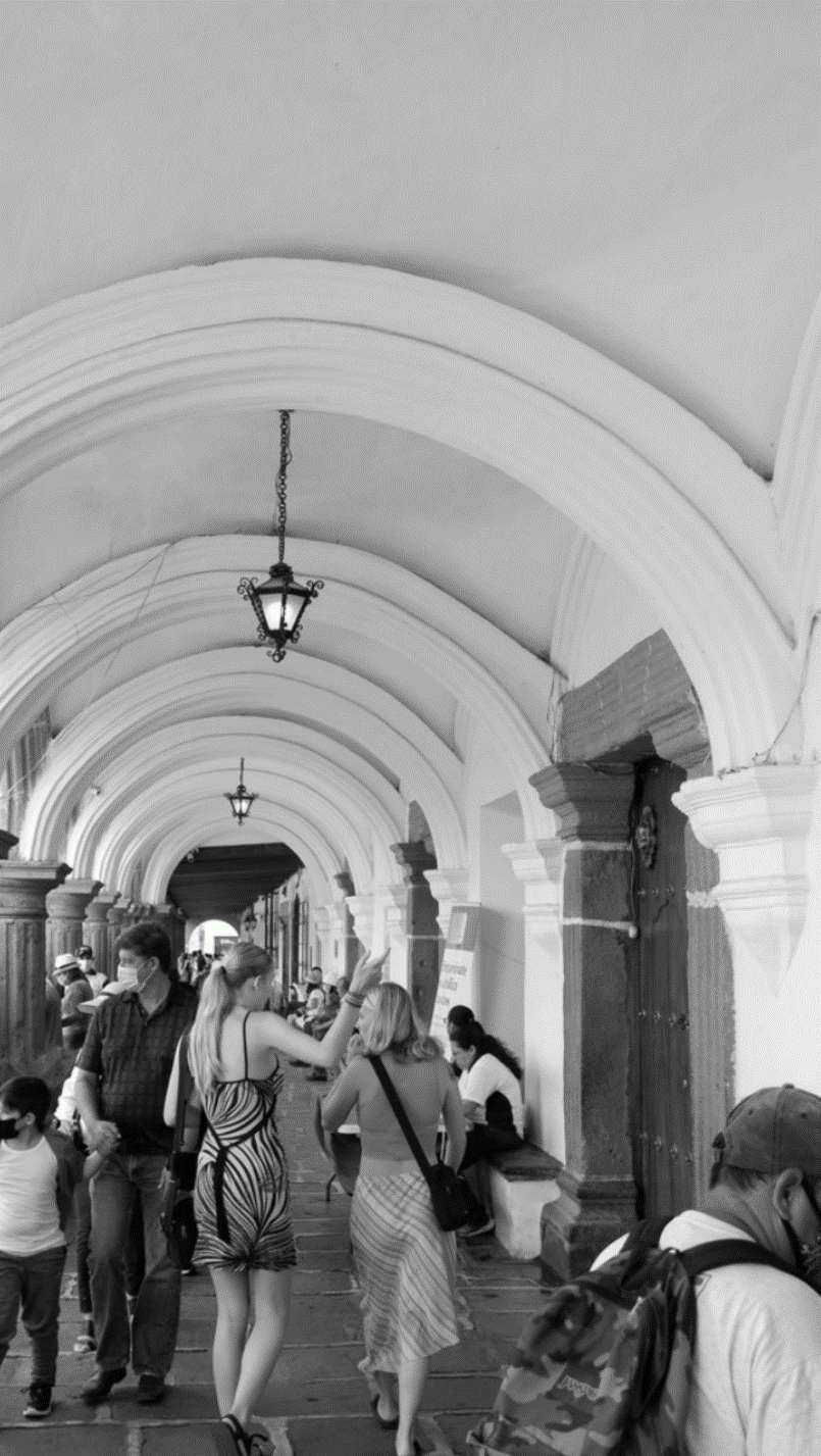People walking through an arched, covered outdoor walkway with hanging lantern lights and wooden columns, some wearing masks.
