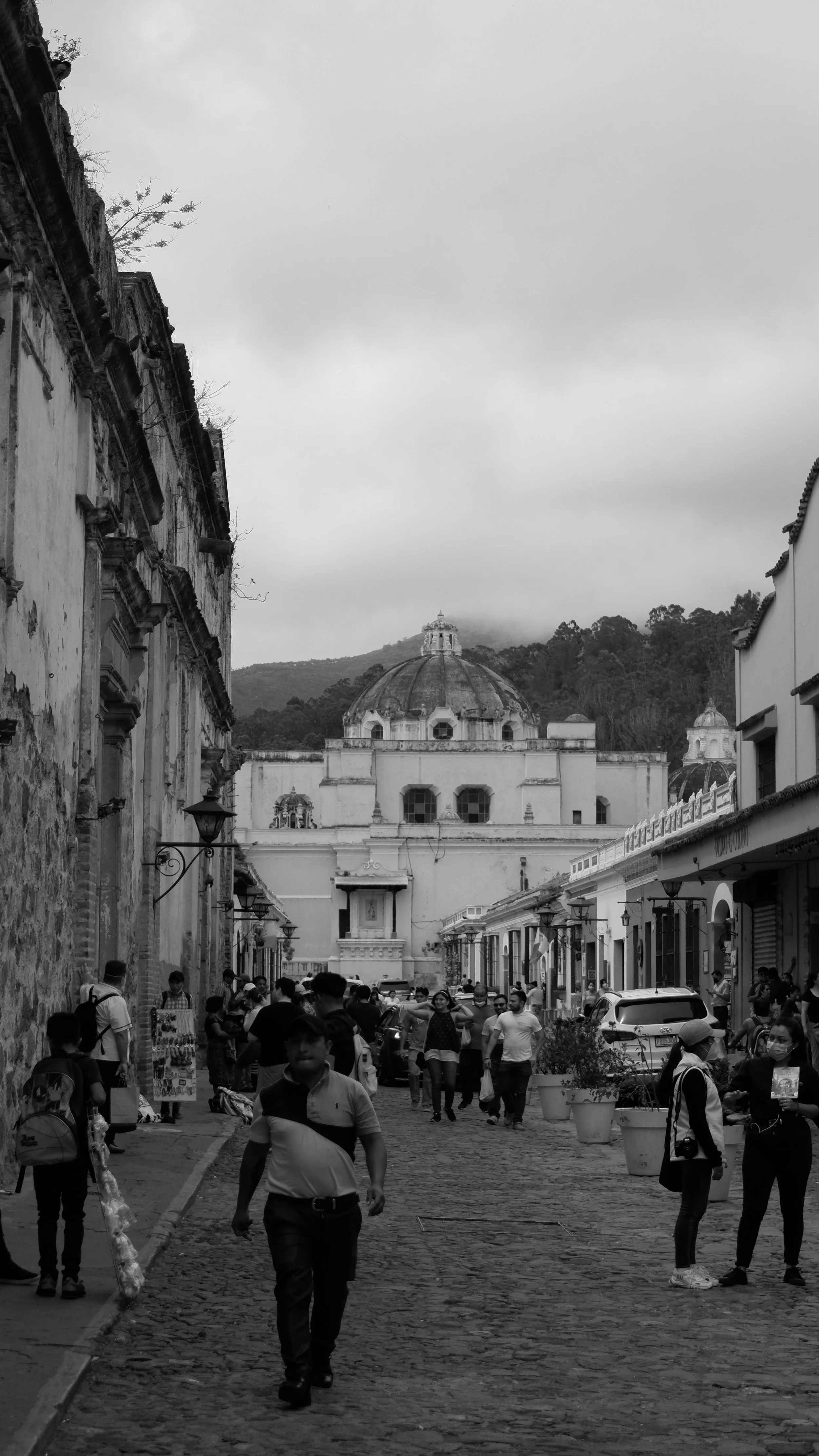A bustling street scene in a historic district with colonial-style buildings on either side, a large domed church in the background, and many people walking and shopping on cobblestone streets.
