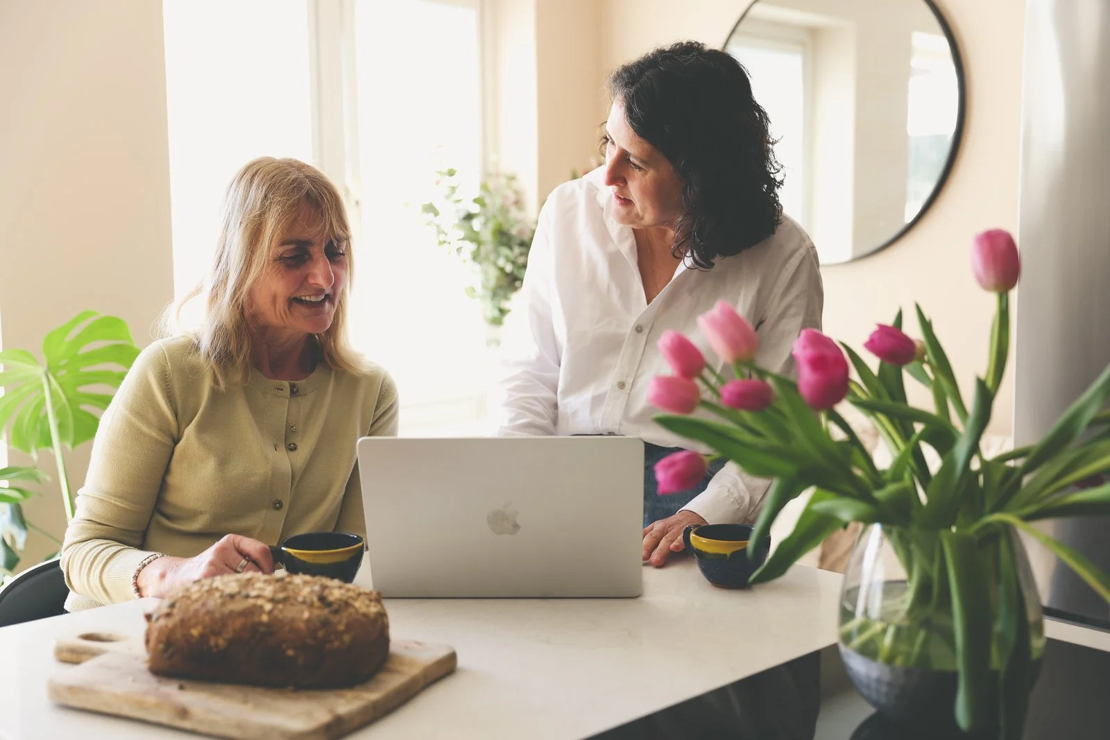 Two midlife women in conversation. Image shows an open laptop, a loaf of bread and coffee cups