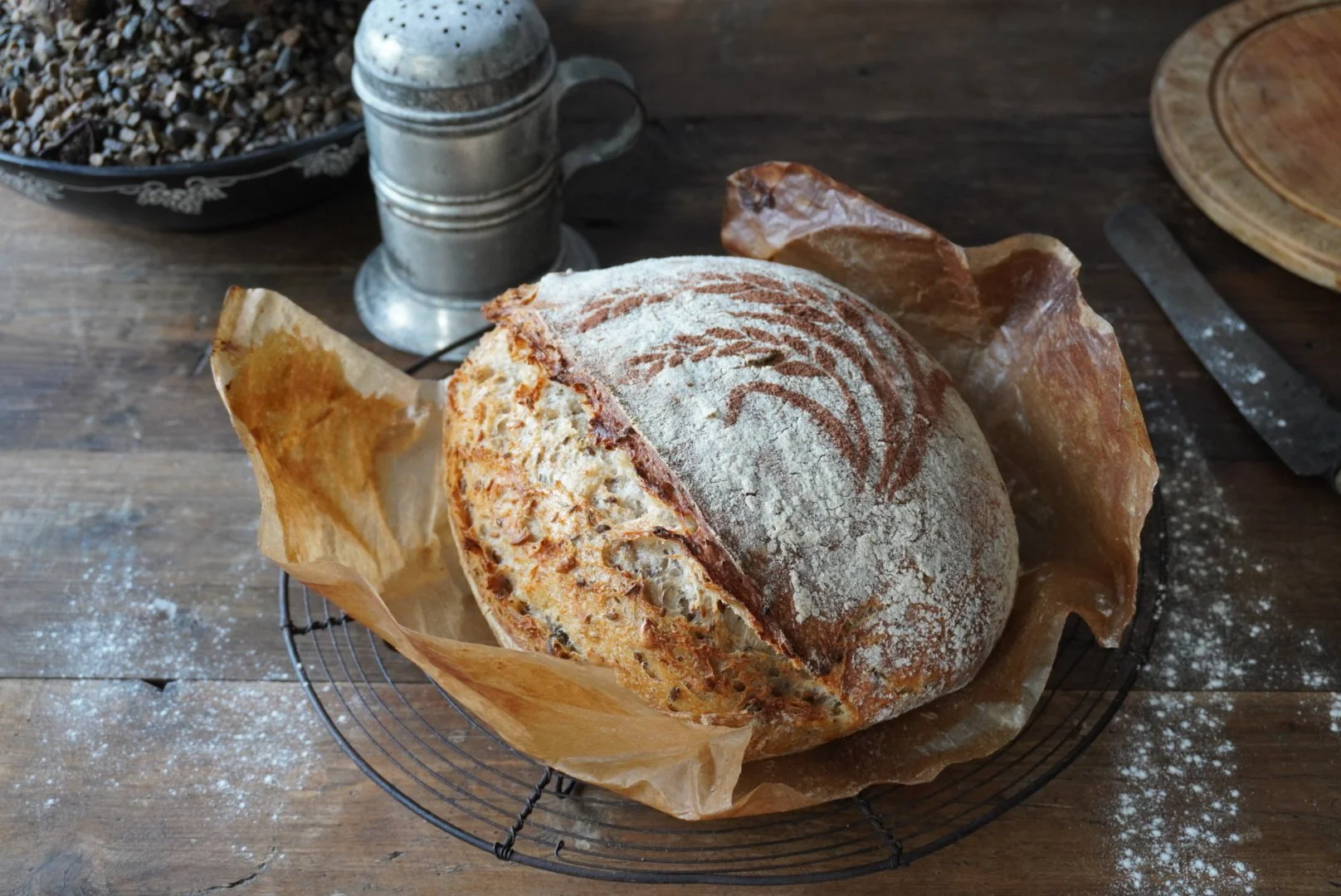 A round loaf of rustic sourdough bread with a flour-dusted crust, resting on brown parchment paper in a wire basket on a wooden table. Behind the bread, there are salt and pepper shakers and a small bowl filled with peppercorns.