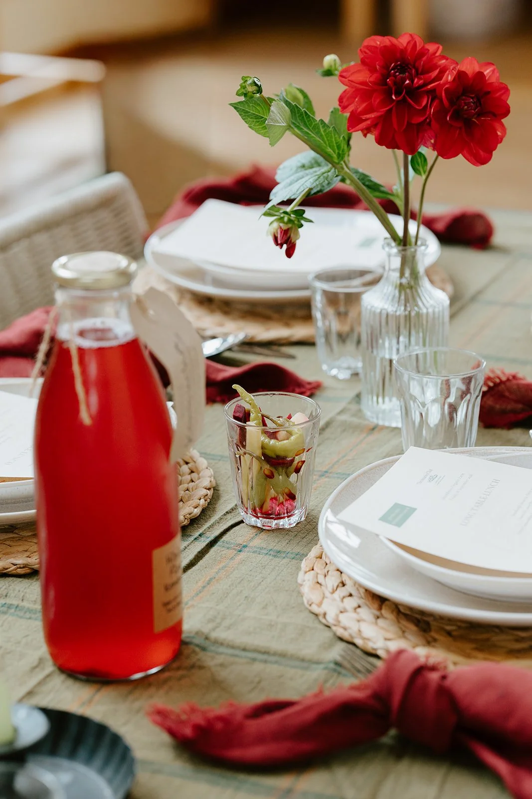 A table set with plates, napkins, a large bottle of red kombucha and a jar of pickles