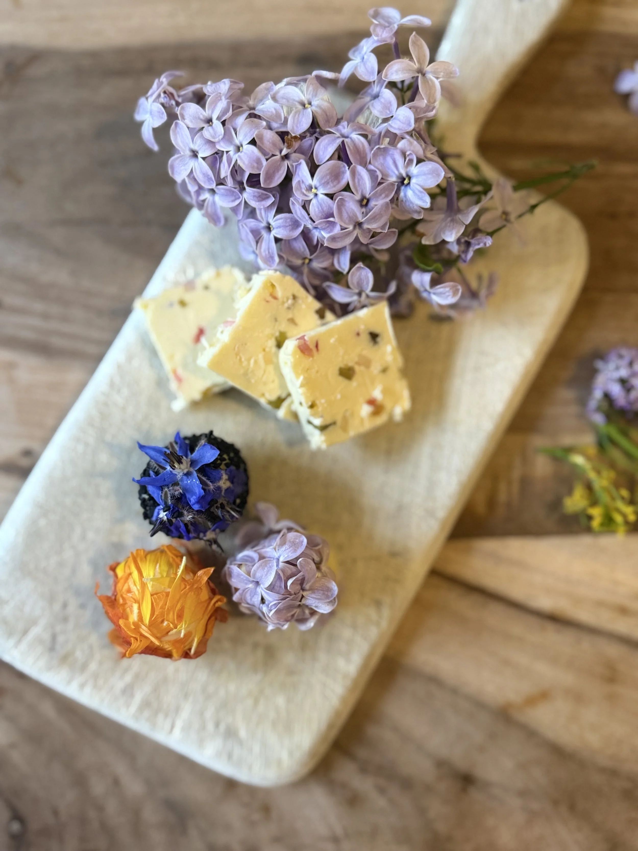 Close-up of a wooden board with purple lilacs, blue cornflower, yellow and orange edible flowers, and pieces of yellow cheese with herbs and red pepper flakes on a light beige surface.