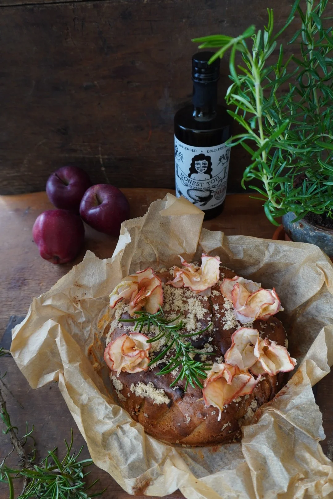 Apple cake decorated with apples and a sprig of rosemary, surrounded by apples, a pot of rosemary and a bottle of 'Honest Toil' olive oil on a wooden surface.