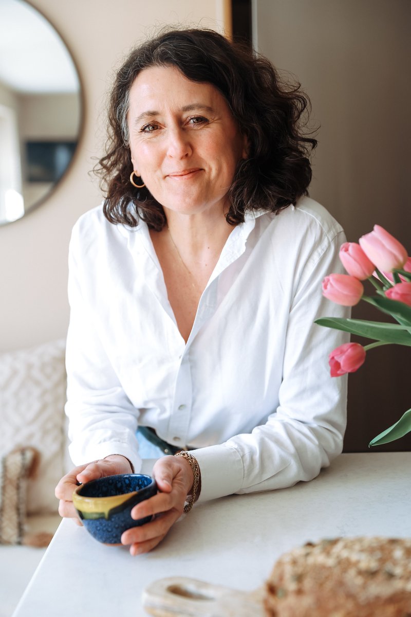 A 50-year-old woman with shoulder-length dark curly hair in a white shirt, smiling softly and holding a blue ceramic cup . There are pink tulips on the right side of the image. In the background, a round mirror hangs on a beige wall.
