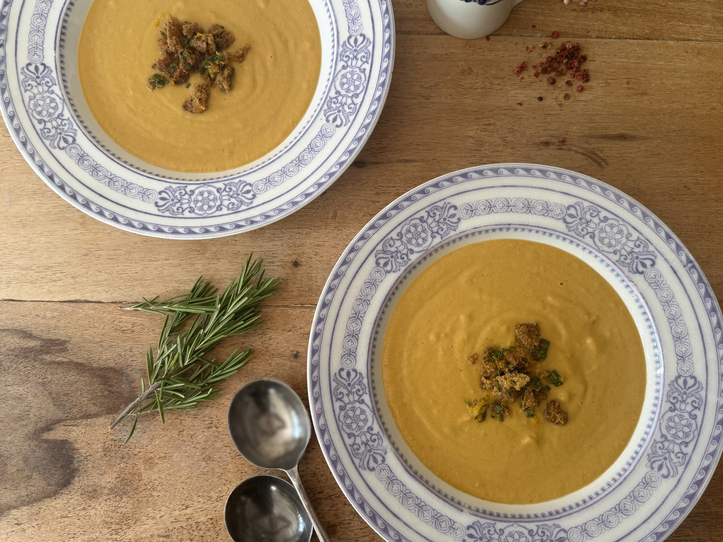 Two bowls of creamy pumpkin soup garnished with chopped herbs, on a wooden table with sprigs of fresh rosemary and two silver spoons nearby.