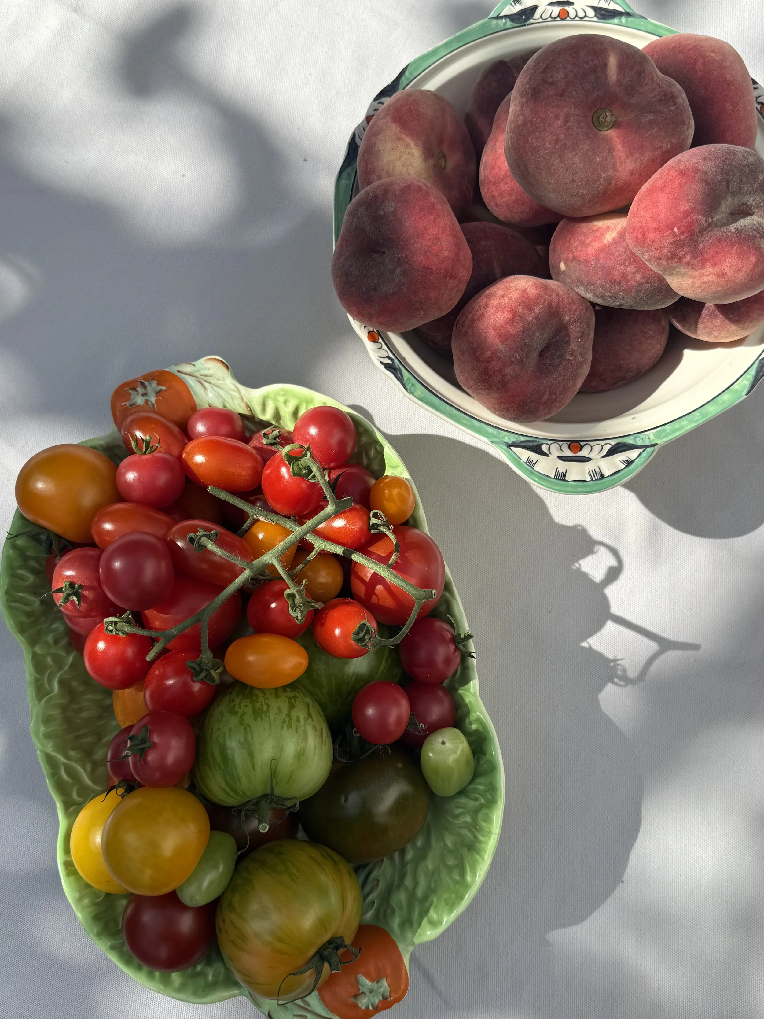 A ceramic bowl filled with peaches, placed on a white surface, with shadows cast by sunlight. Next to it, a decorative green dish holds an assortment of different coloured tomatoes and a green tomato.