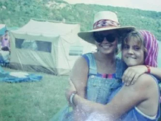 Two women hugging outdoors at a camping site, with tents and green trees in the background.