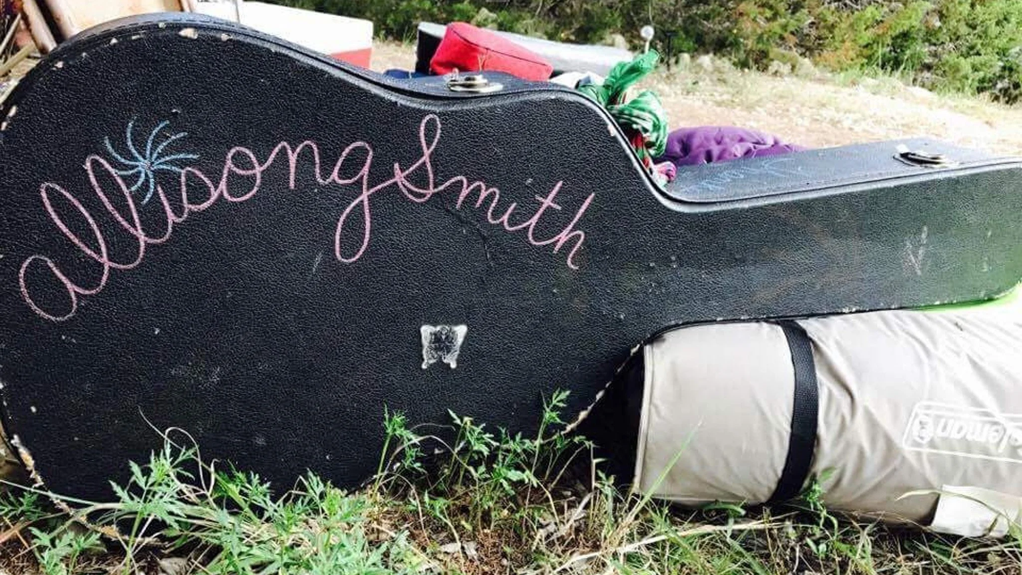 Allisong Smith's guitar - A black guitar case with a pink name "Allisong Smith" written on it. The case is placed on the ground among grass and small plants, with some camping or outdoor gear nearby.