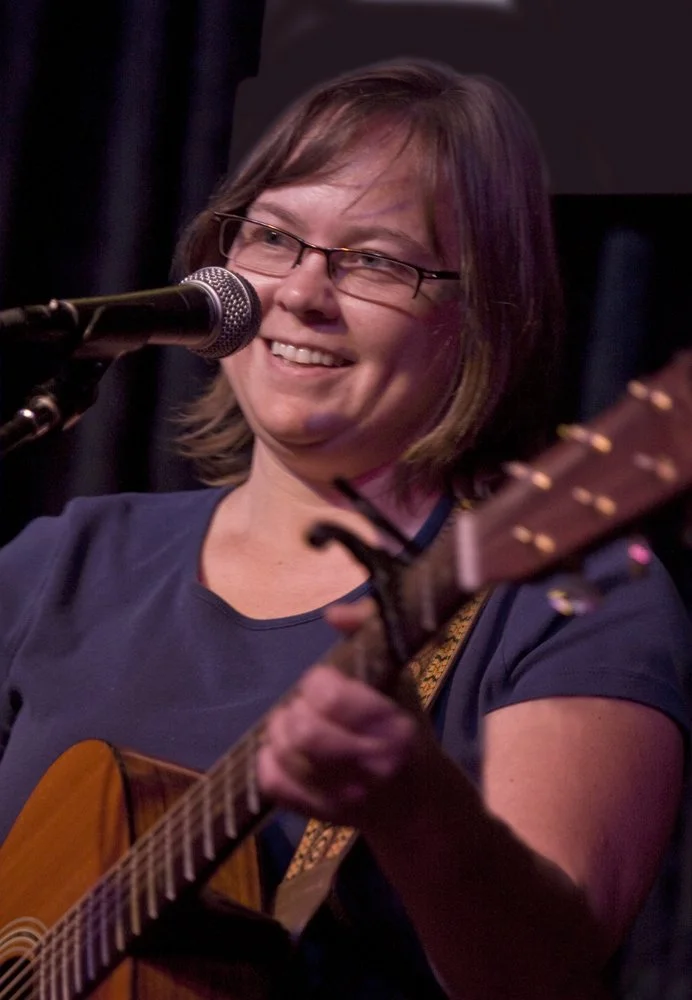 Woman with glasses smiling and singing into a microphone while playing an acoustic guitar.