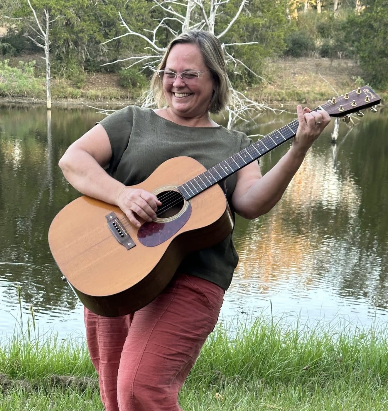 A woman smiling and playing an acoustic guitar outdoors near a body of water with trees in the background.
