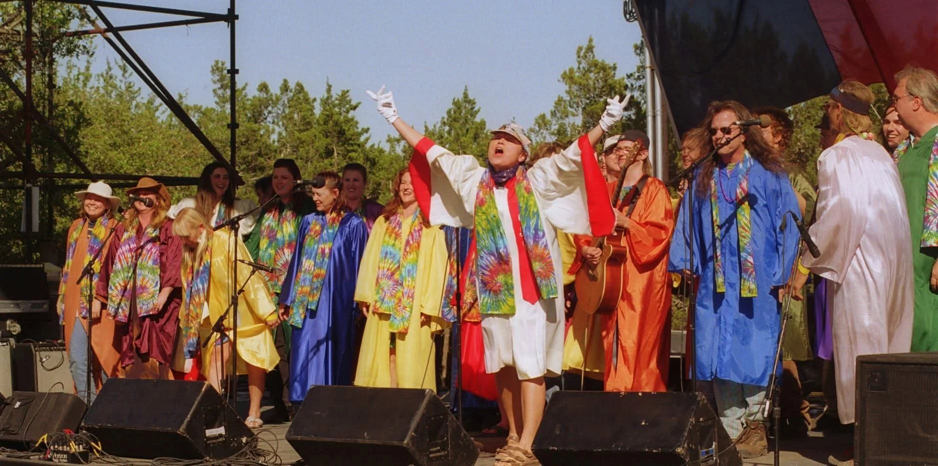 A group of people dressed in colorful tie-dye and rainbow robes performing on an outdoor stage, some holding microphones and a guitar, with a woman at the center raising her arms in celebration.