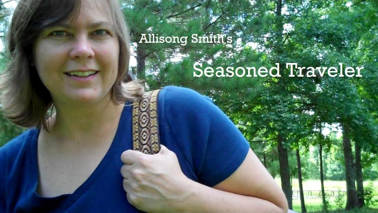 Allisong Smith's Seasoned Traveler - A woman with short brown hair smiling outdoors, carrying a guitar with a patterned strap, and surrounded by green trees.