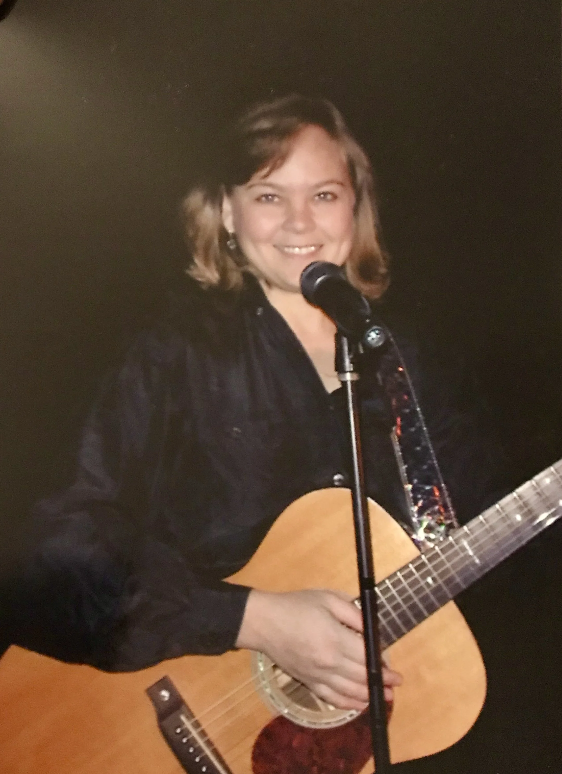 Allison Smith - Person playing an acoustic guitar and singing into a microphone, wearing a black shirt, with a dark background.