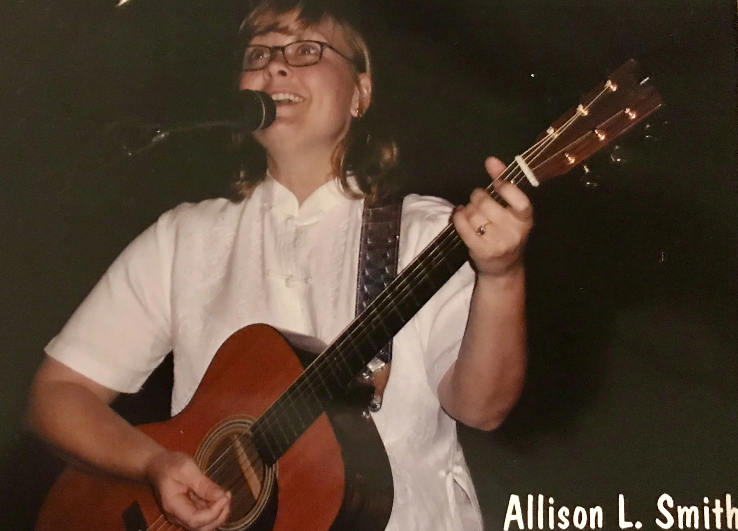 A woman with glasses singing into a microphone while playing an acoustic guitar, wearing a white shirt, with text 'Allison L. Smith' at the bottom.